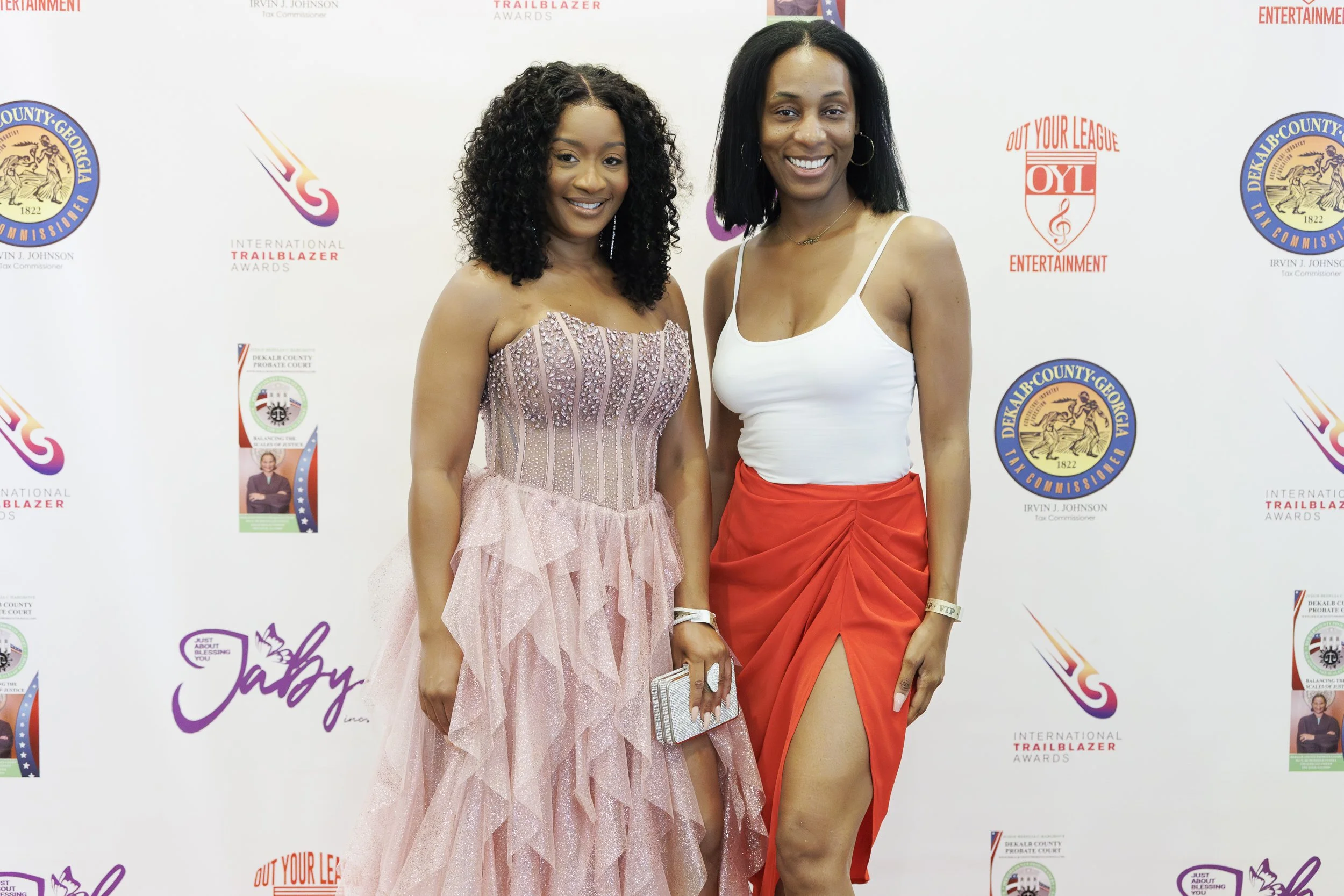 Two women smiling and posing together at an event, one in a pink strapless gown with ruffles and the other in a white tank top and red skirt with a slit, standing in front of a backdrop with various logos.