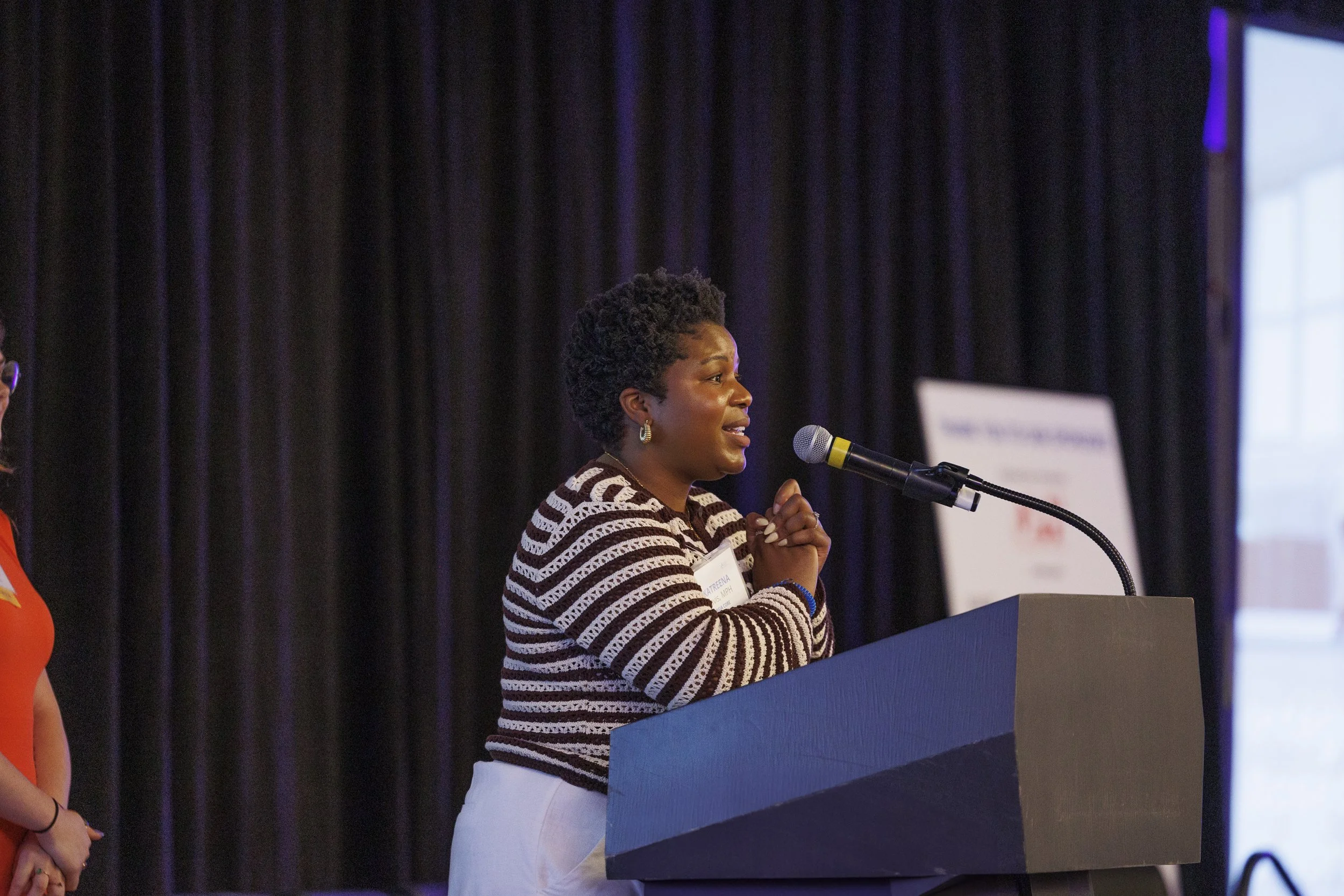Woman at a podium speaking into a microphone during a conference or event, with black curtains in the background.