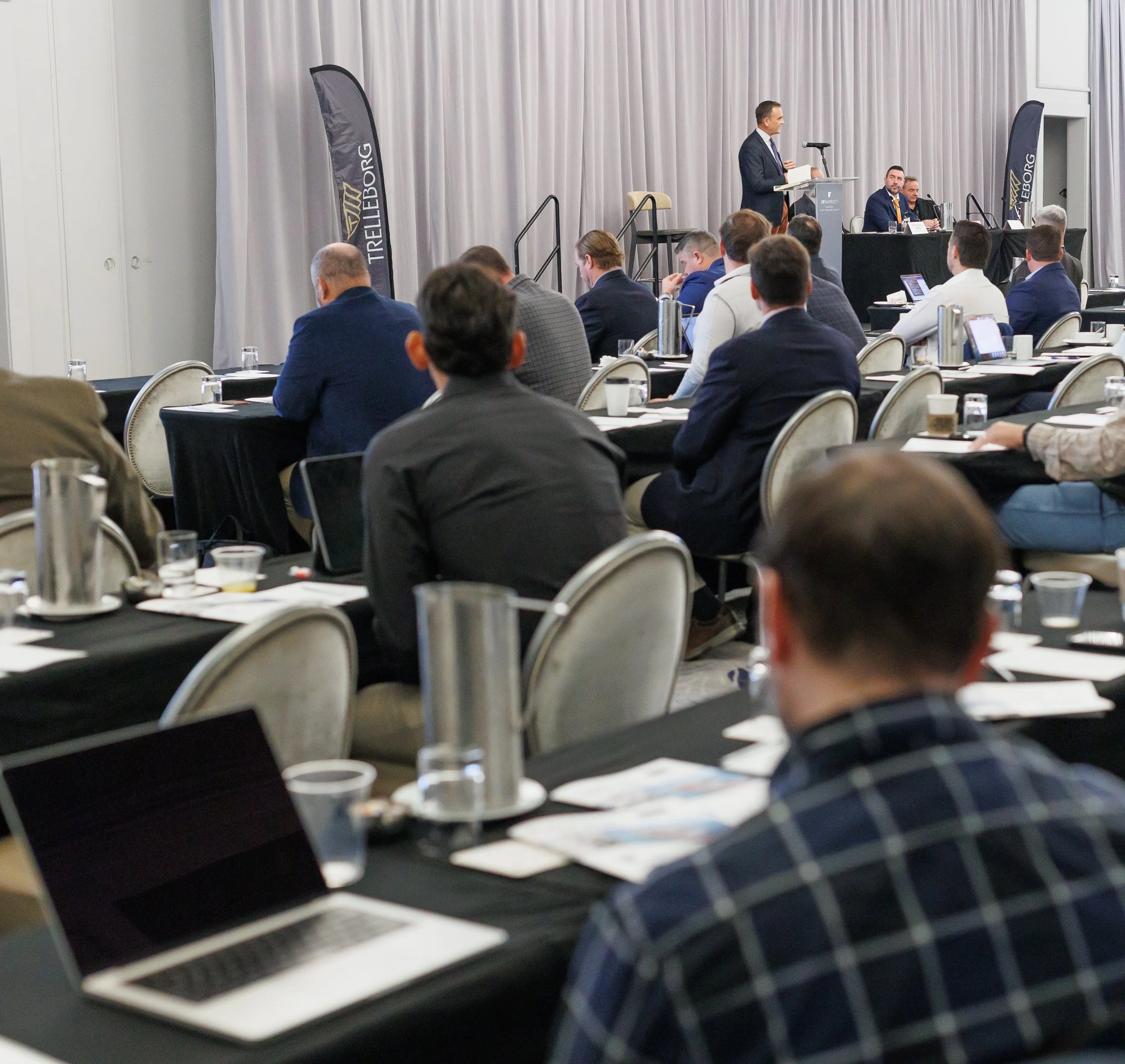 Business conference with attendees seated at tables, listening to a speaker at a podium on stage with banners behind, in a hotel conference room.