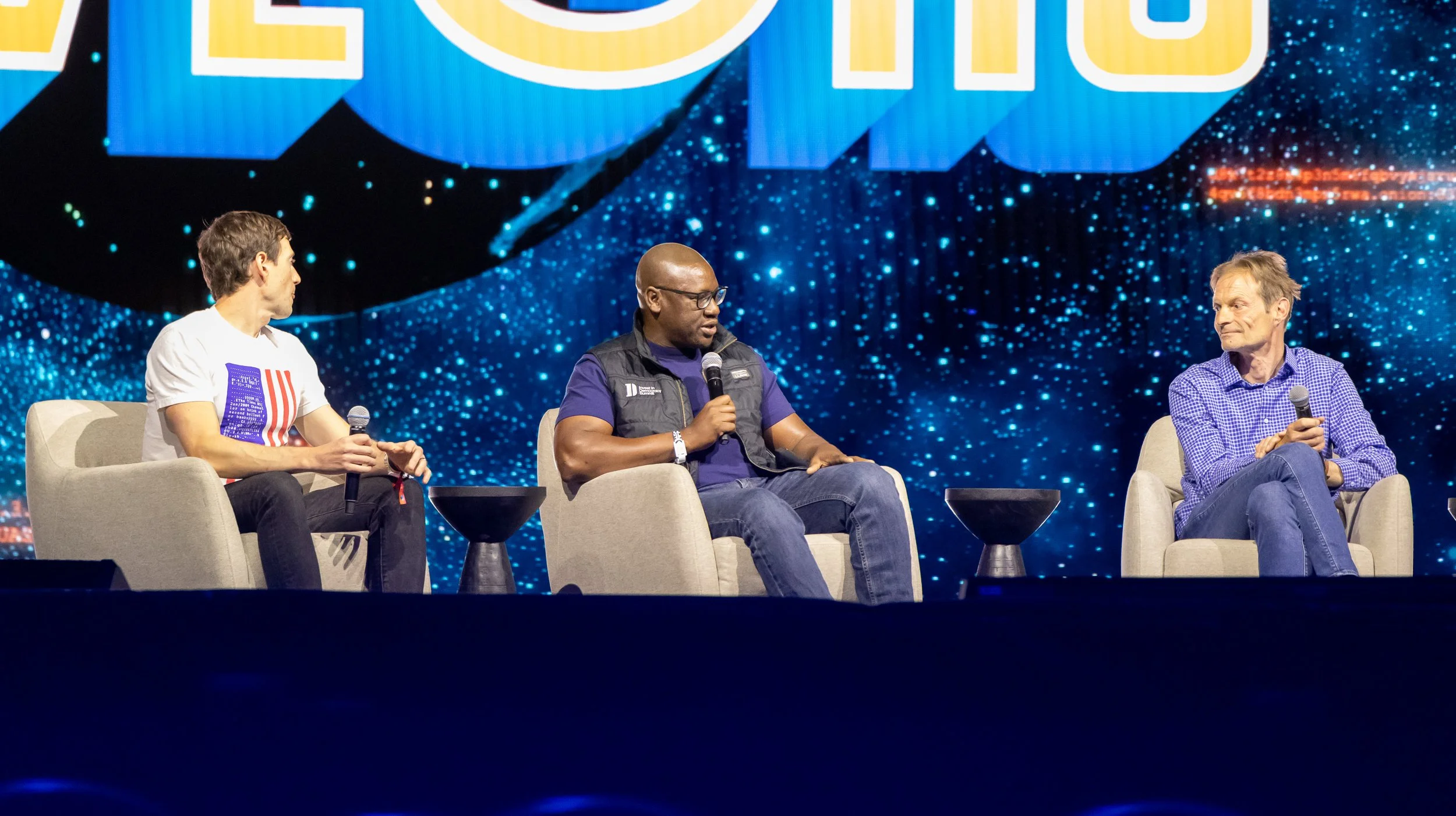 Three men sitting on stage in armchairs holding microphones during a panel discussion at a conference, with a digital background display.