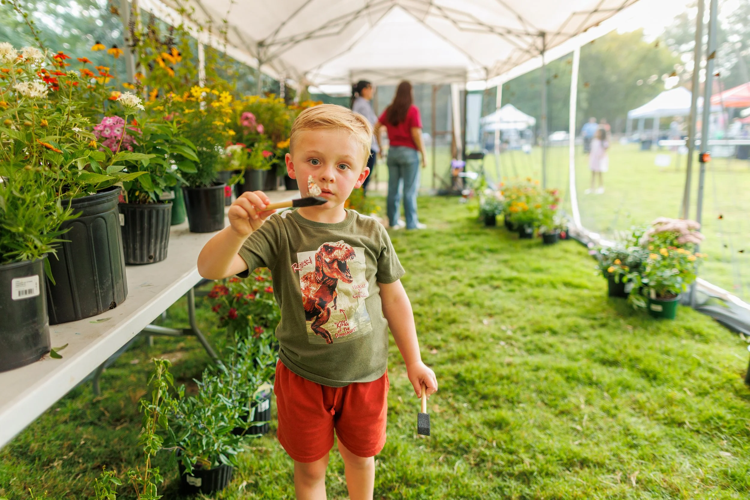 A young boy with blonde hair holding a gardening tool, standing in front of potted flowers at an outdoor plant market or fair, with tents and other people in the background.