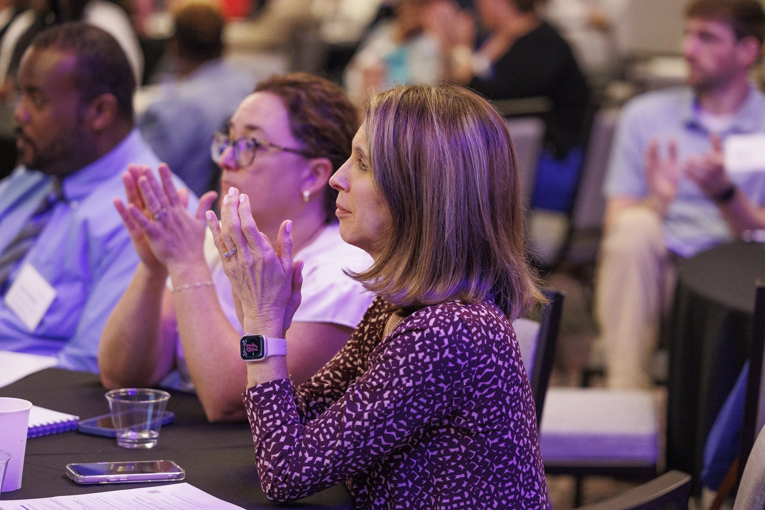 A woman with brown hair wearing a patterned purple top, sitting at a conference table, clapping. She has a smartwatch on her wrist, with papers, a phone, and a cup in front of her. Other attendees are seated around, some also clapping.