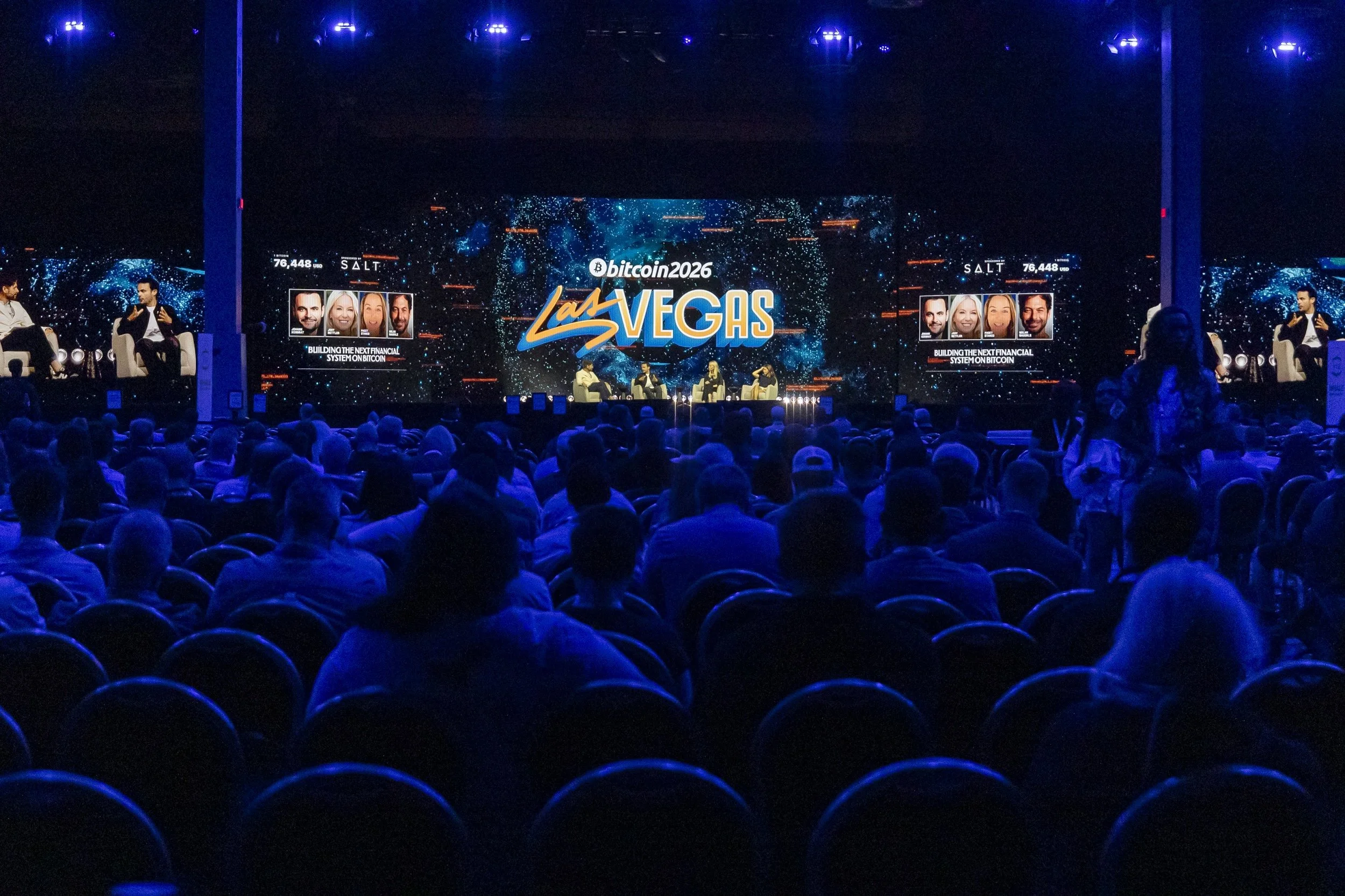 An indoor conference or event center with a large audience seated and a stage with a big digital screen displaying the words 'Bitcoin 2026 Las Vegas' and images of four speakers.