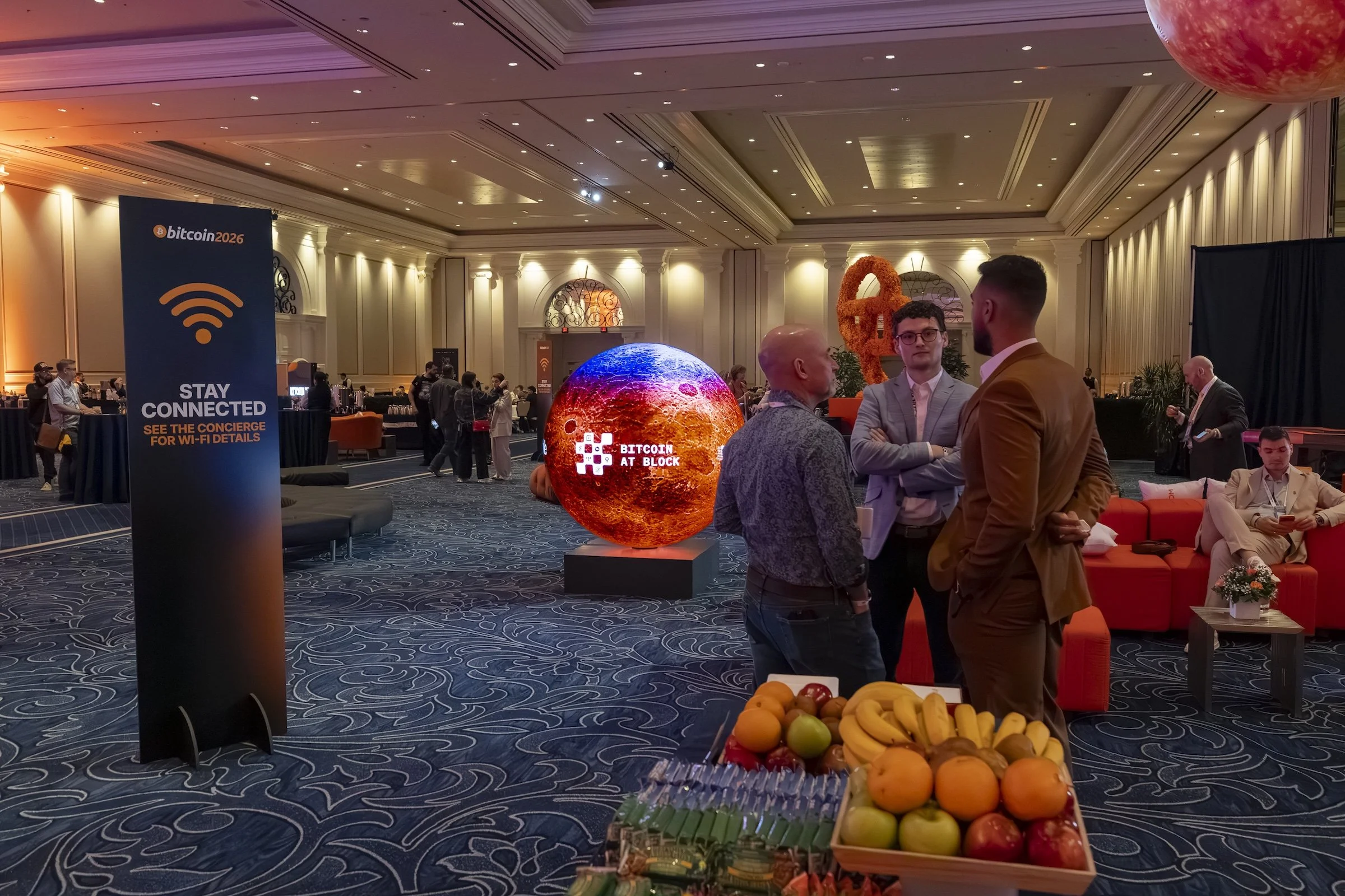 An indoor conference or convention setting with people gathered around. There is a large glowing globe with the text 'Bitcoin at Block' and a sign that reads 'Bitcoin 2026' indicating a Bitcoin or cryptocurrency event. The room has elegant decor with