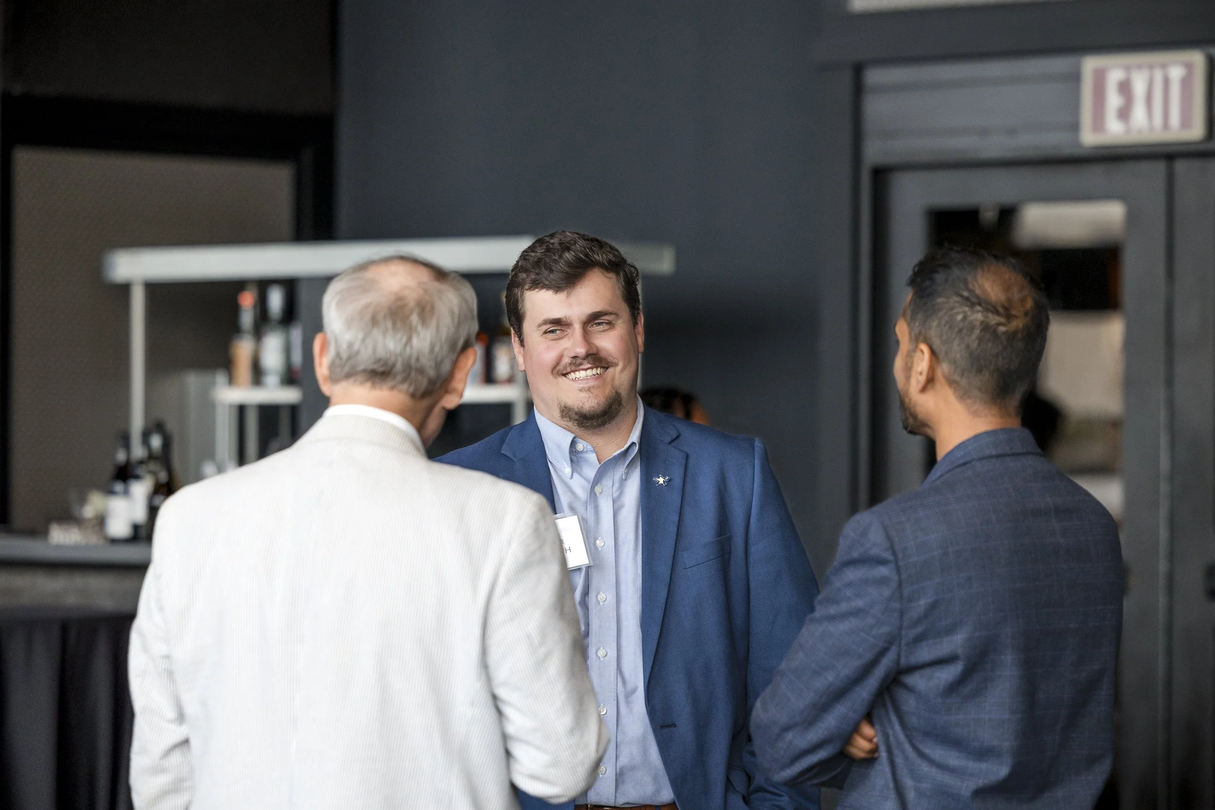 Three men in business attire engaging in conversation at a professional event indoors, with a bar area and exit sign in the background.