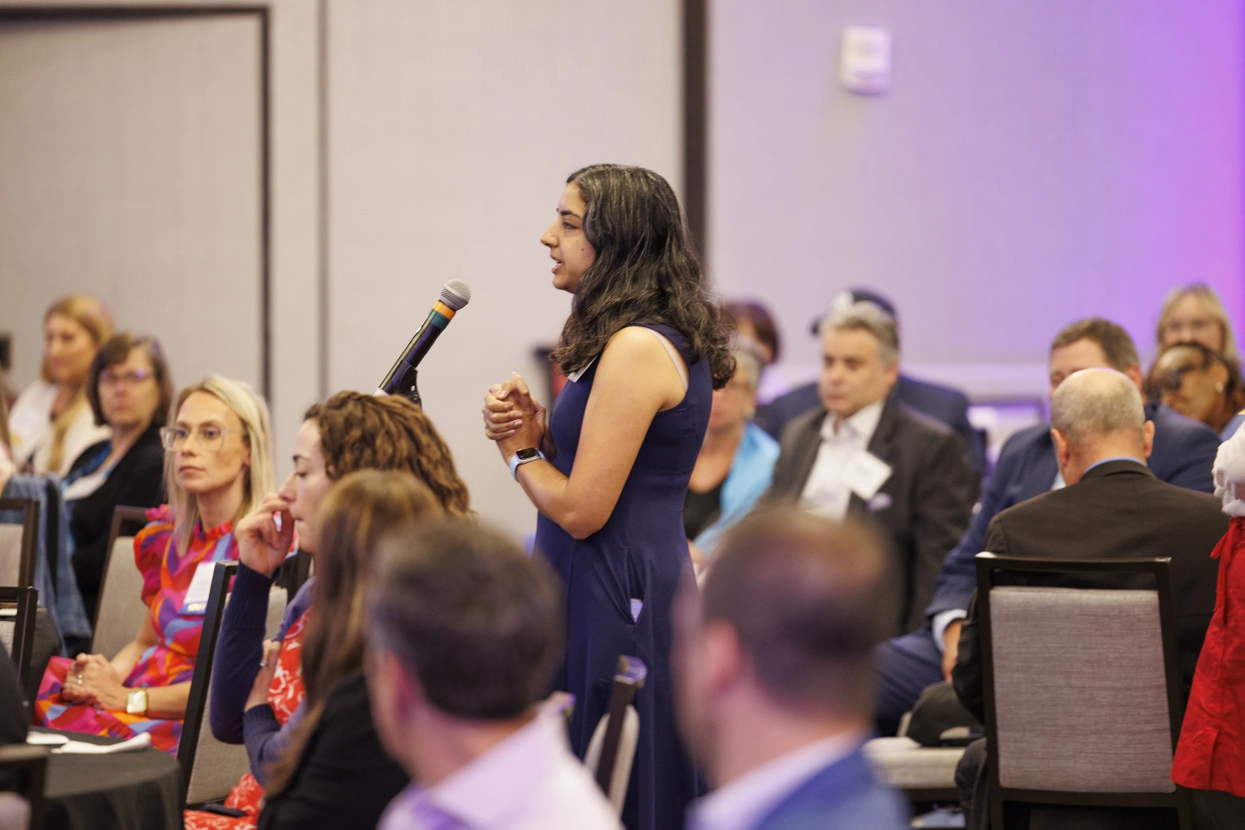 A woman in a blue dress speaking into a microphone during a conference or seminar, with seated attendees listening.