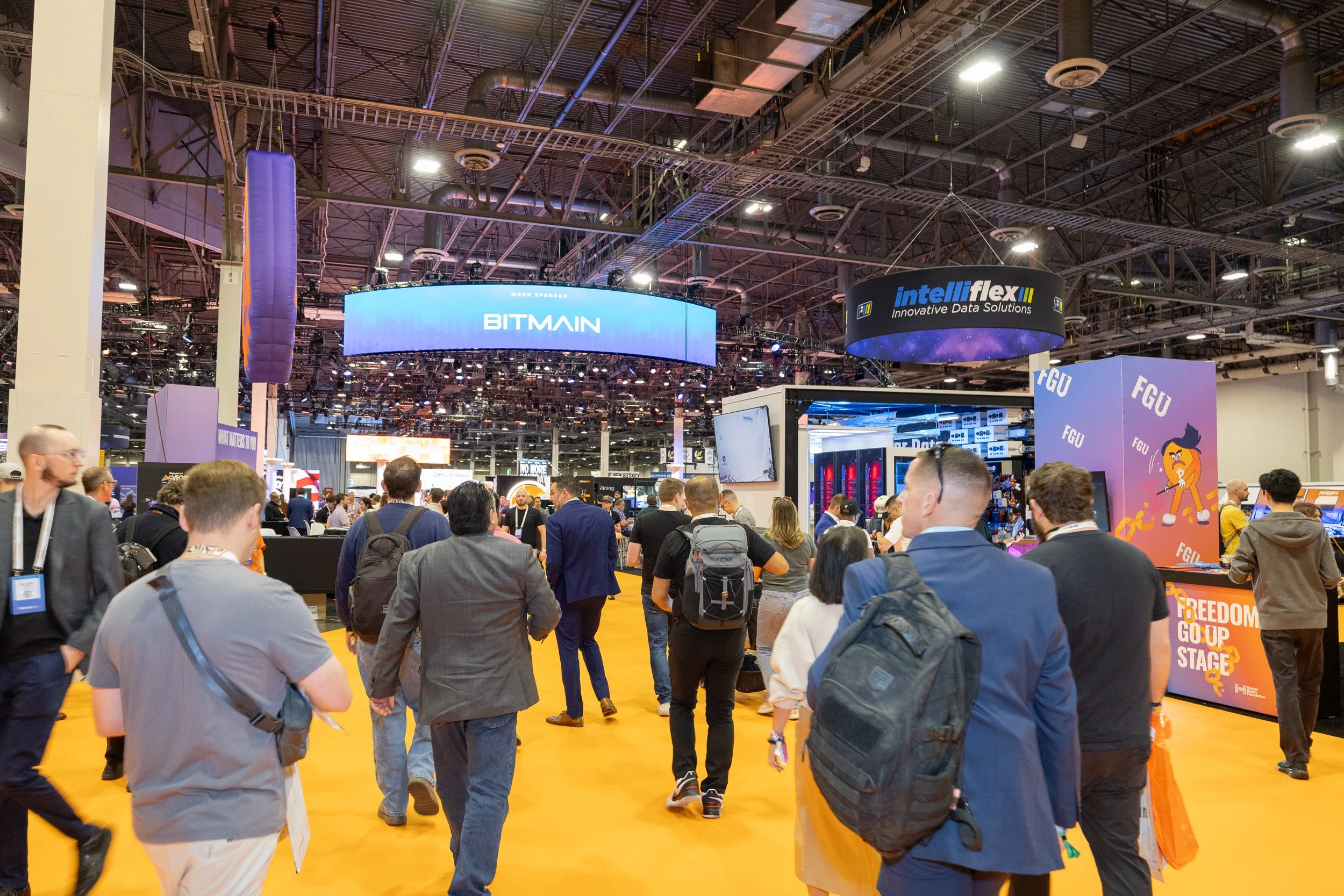 A crowded industry conference with numerous booths and large digital signs overhead displaying company names such as BITMAIN and intelliflex. Attendees are walking and standing around, engaging with exhibits across a yellow carpeted floor in a large 