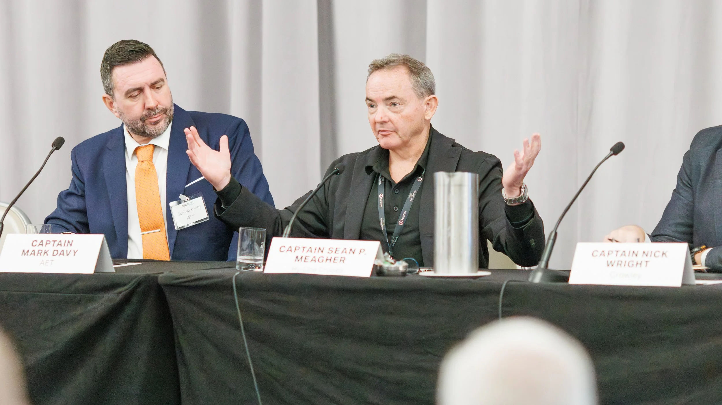 Three men sitting at a panel discussion table with name tags; the man in the middle gestures with his hands while speaking.