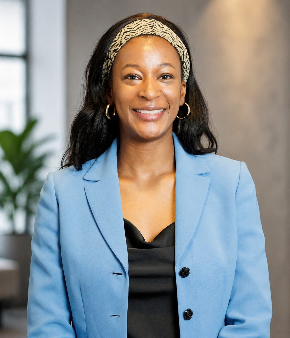 A smiling woman with dark hair wearing a headband, gold hoop earrings, a blue blazer, and a black top, standing in an office environment corporate professional headshot