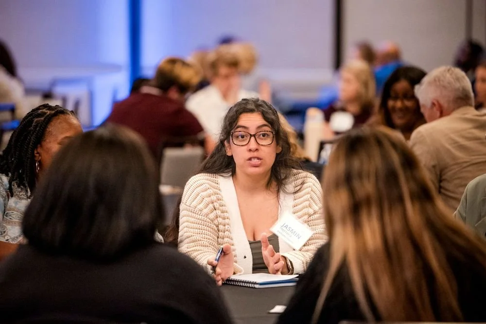A woman with glasses and long dark hair, speaking at a roundtable during a conference, with other attendees in the background.