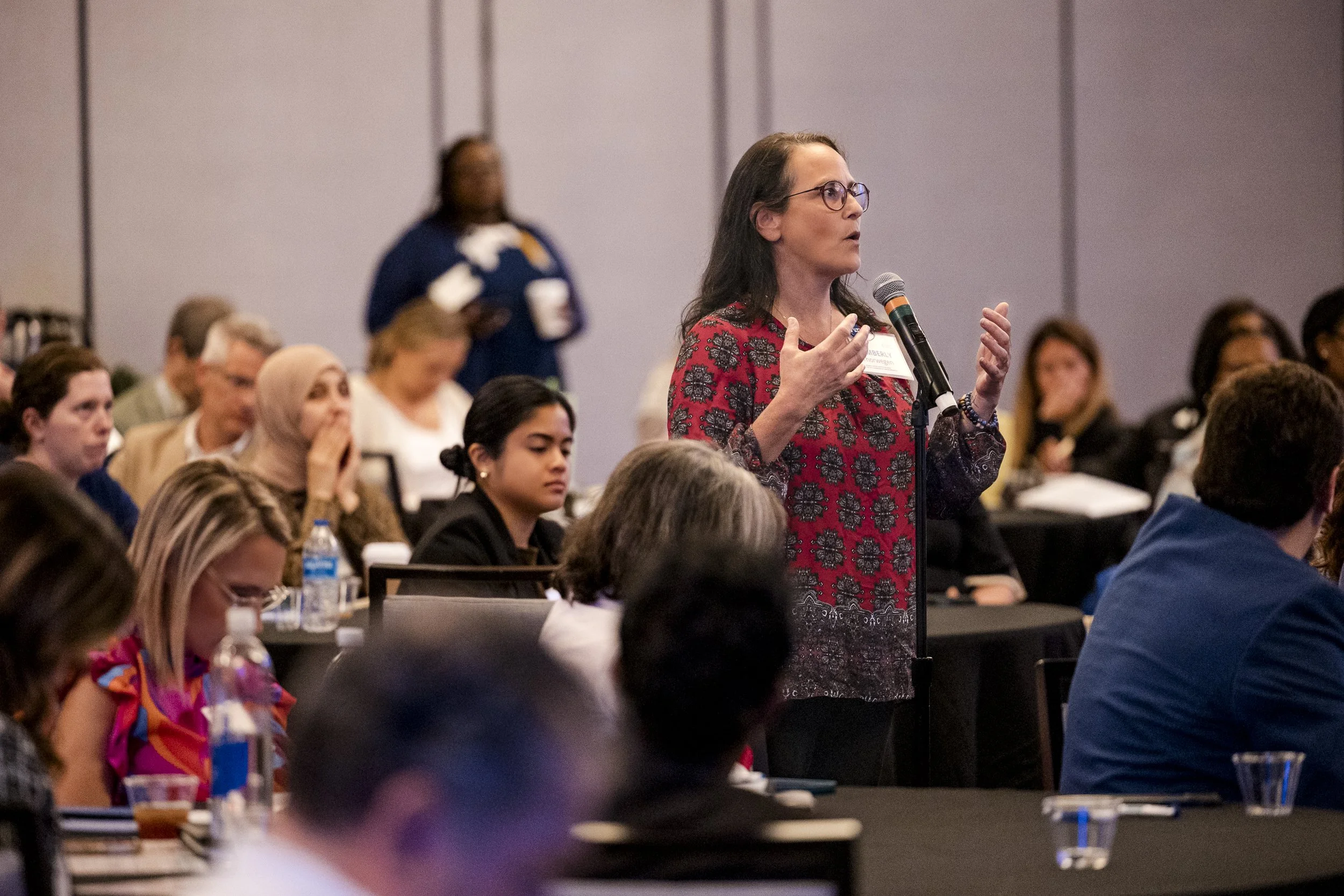 A woman with glasses and long dark hair, wearing a red and black patterned top, is speaking into a microphone at a conference or seminar. She is standing among seated attendees who are listening attentively, with notebooks and water bottles on the ta