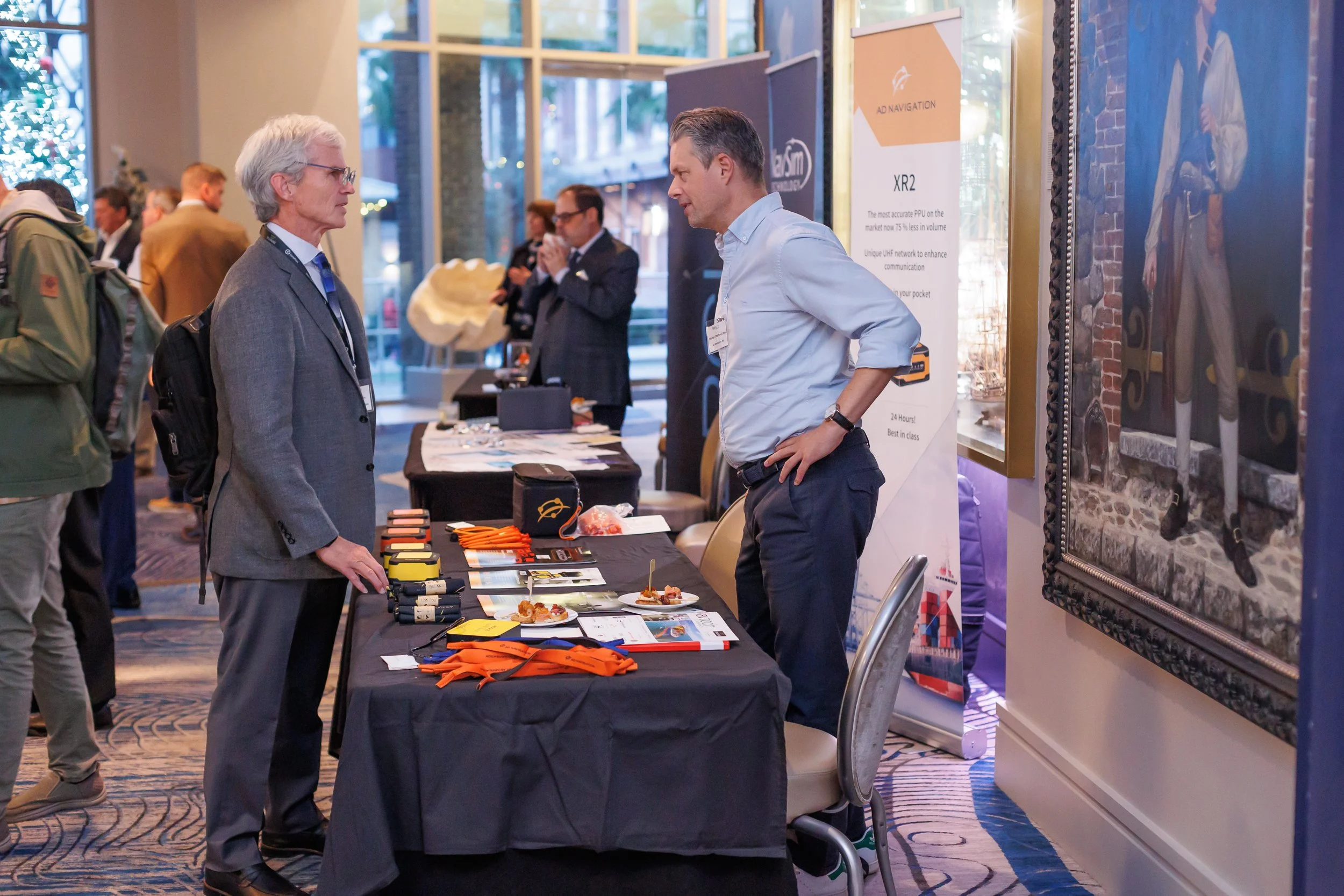 At a conference, a man in a gray suit with glasses is talking to a young man in a light blue shirt behind a booth displaying promotional items, with other conference attendees in the background.