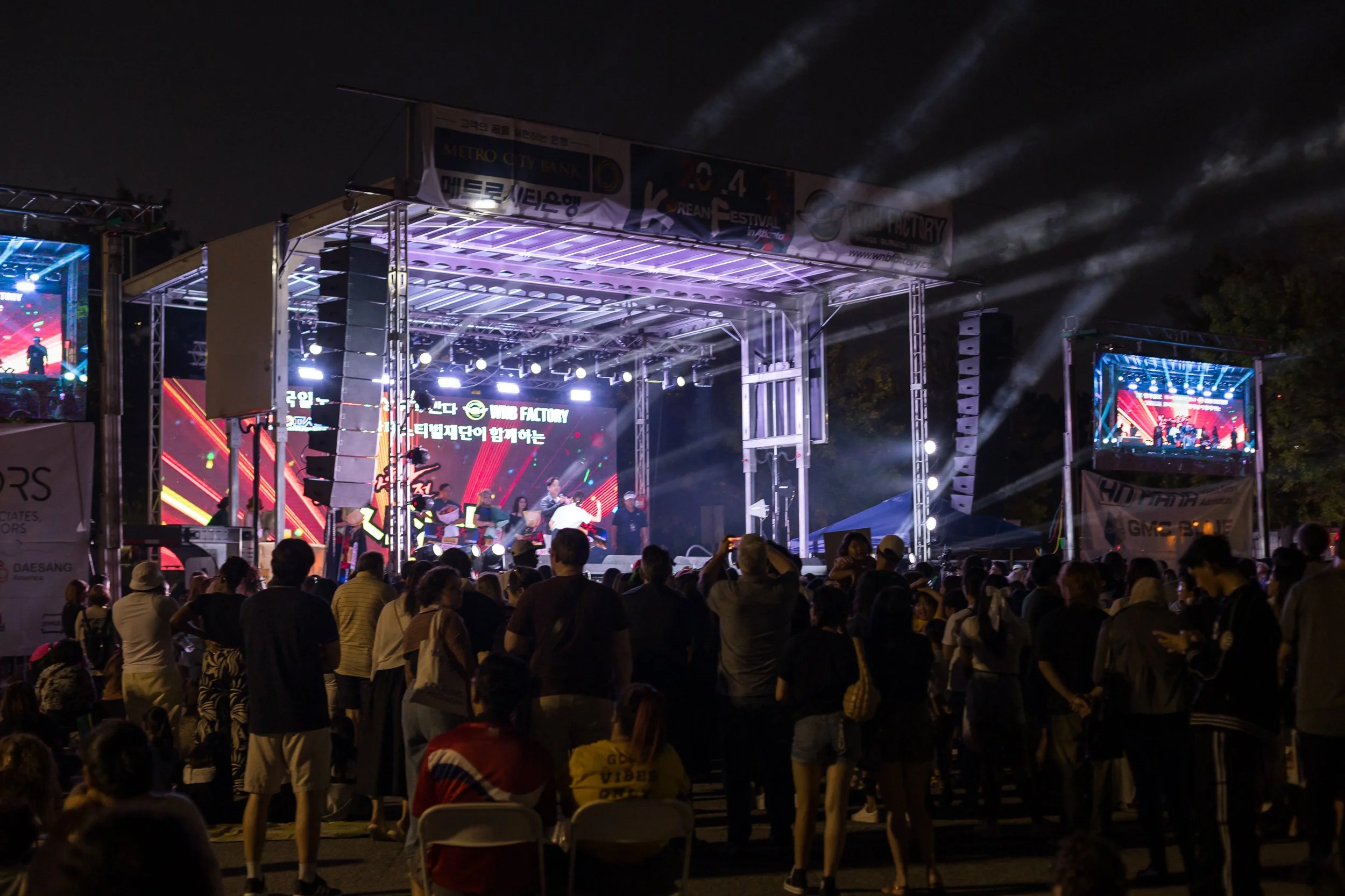Nighttime outdoor concert with a large crowd standing and sitting, stage with bright lights and screens, smoke effects, and trees in the background.