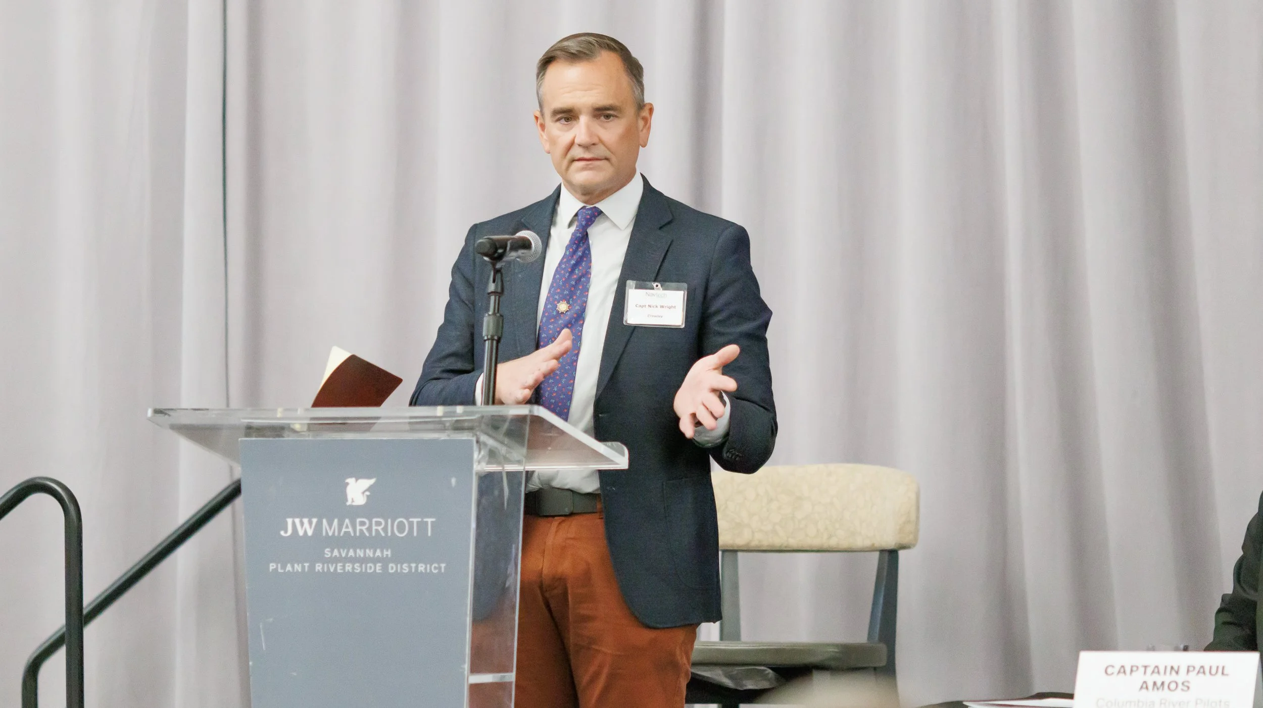 A man in a suit and tie standing at a podium with a microphone, giving a presentation at JW Marriott Savannah Plant Riverside District. He has short, neatly combed hair and is wearing a name tag. There are chairs and a table with a nameplate in front