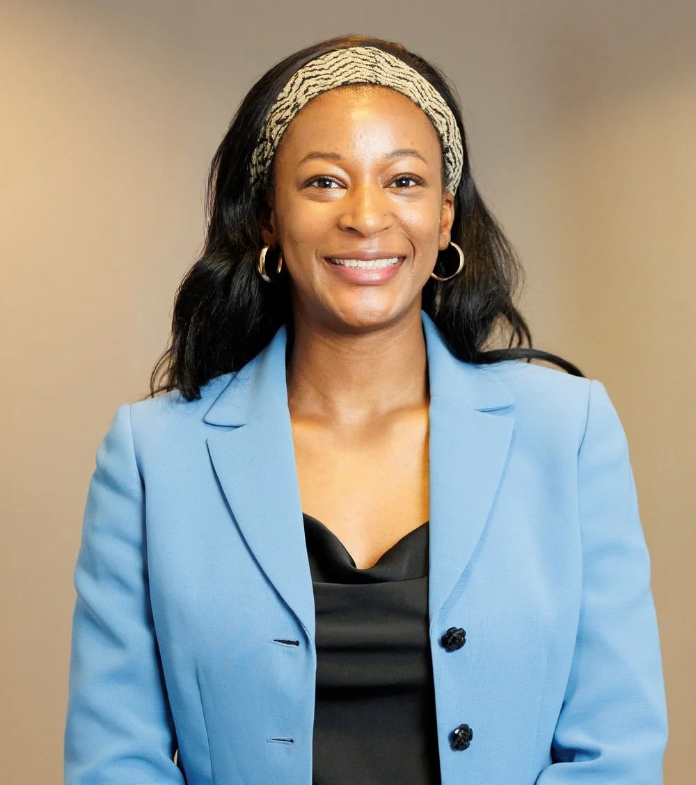 A smiling woman with dark, wavy hair wearing a blue blazer, black top, hoop earrings, and a patterned headband against a neutral background. For corporate headshots in metro Atlanta.