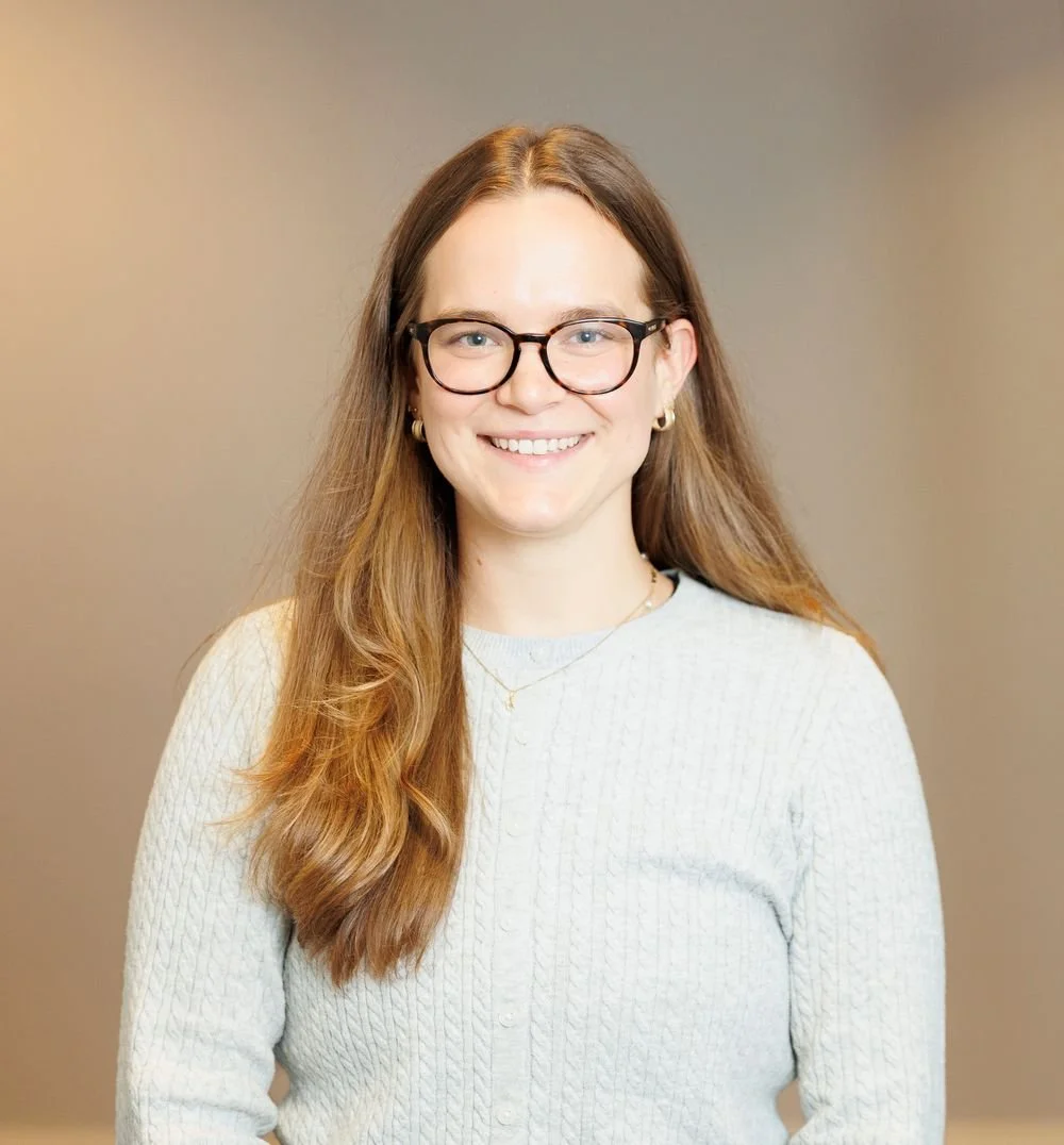 A young woman with long red hair, glasses, and gold earrings smiling at the camera against a plain background.