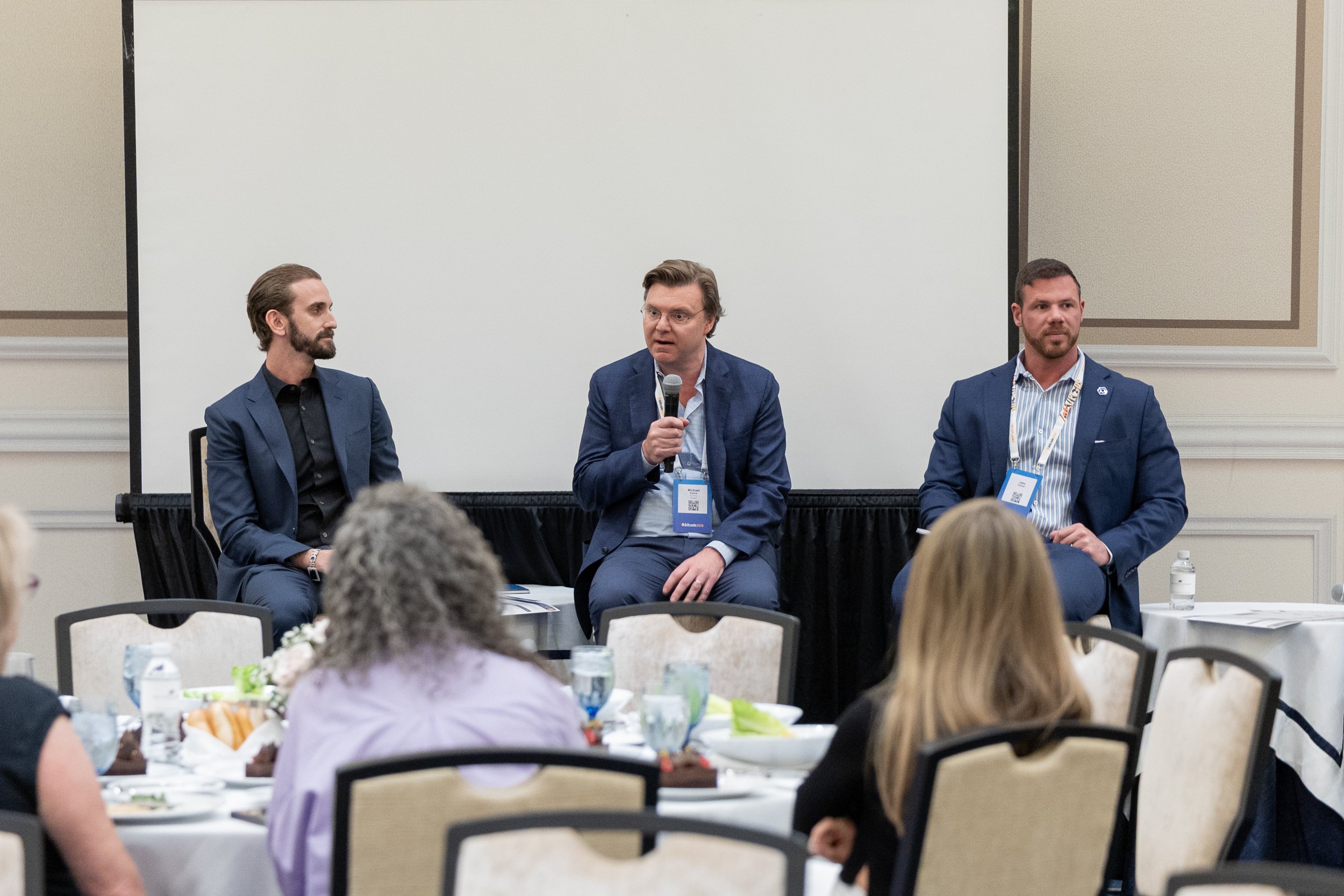 A panel discussion at a conference with three men seated at the front, one of whom is speaking into a microphone, while the audience and a decorated table are visible in the foreground.
