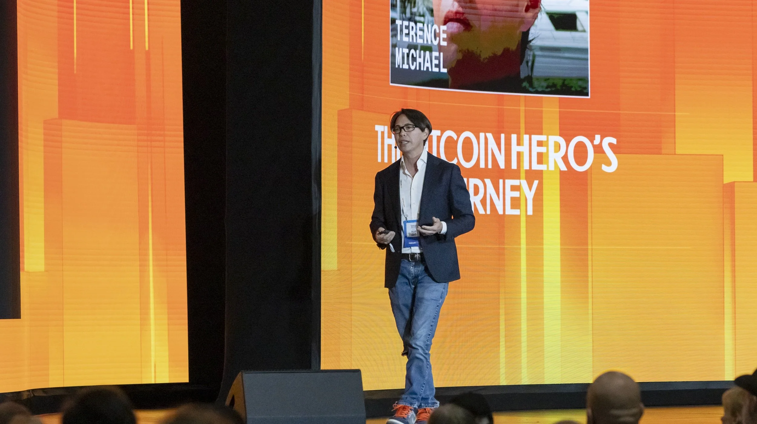 A woman with short dark hair and glasses presenting on stage during a conference. She is wearing a navy blazer, white shirt, and jeans, holding a remote control. Behind her is a large orange screen displaying the text "The Bitcoin Hero's Journey" and