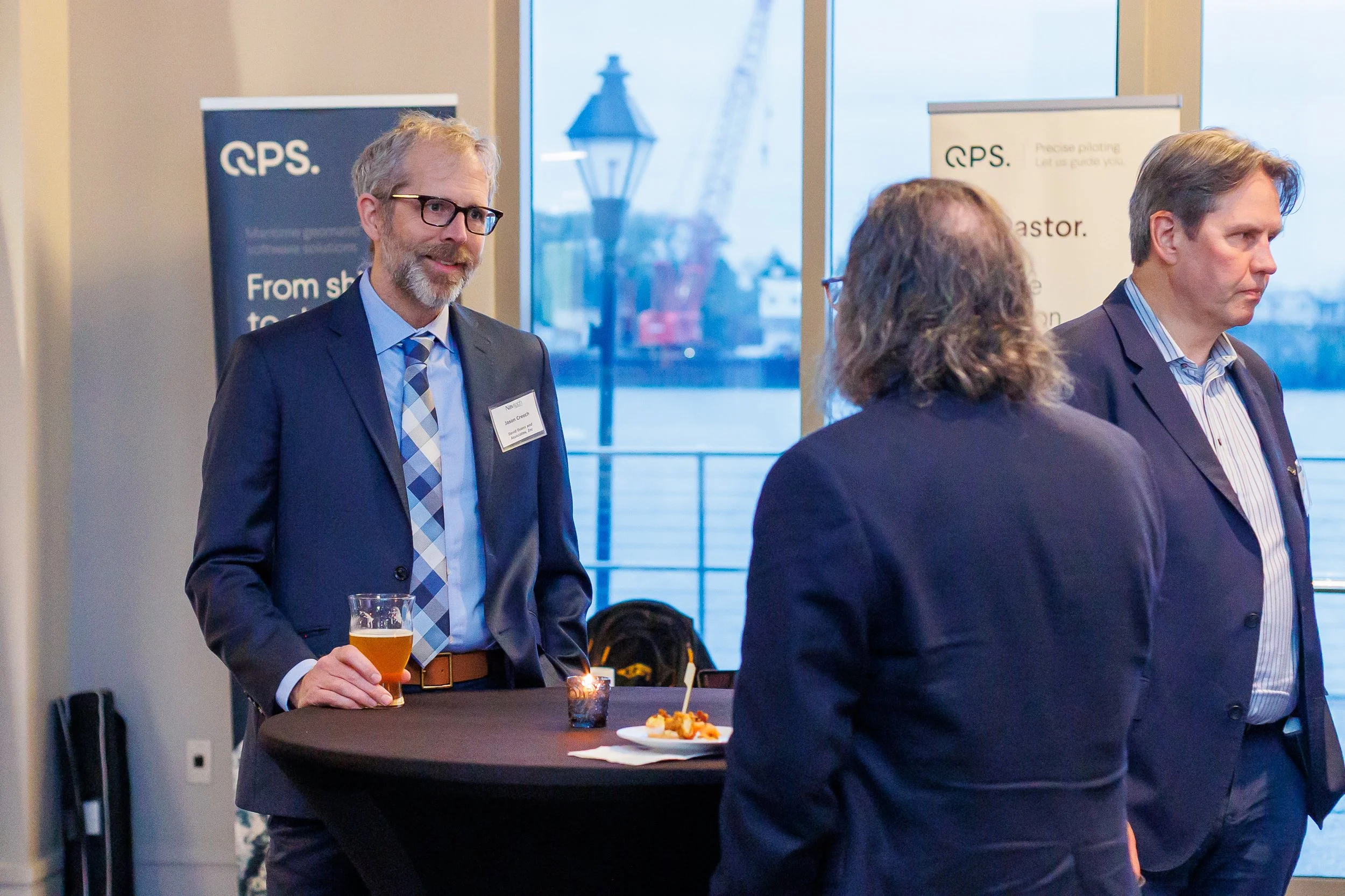 Three people in business attire having a conversation at a networking event with drinks and snacks, with banners and water outside in the background.