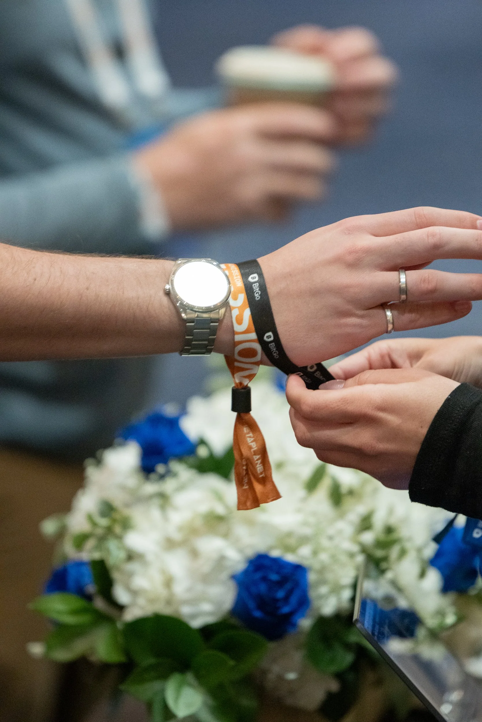 A person wearing a watch and a wristband with the text 'Bego' on it is holding a small white dish with pink-colored food on it. Another person is reaching out with their hand, wearing a ring. There are white flowers with blue accents in the backgroun