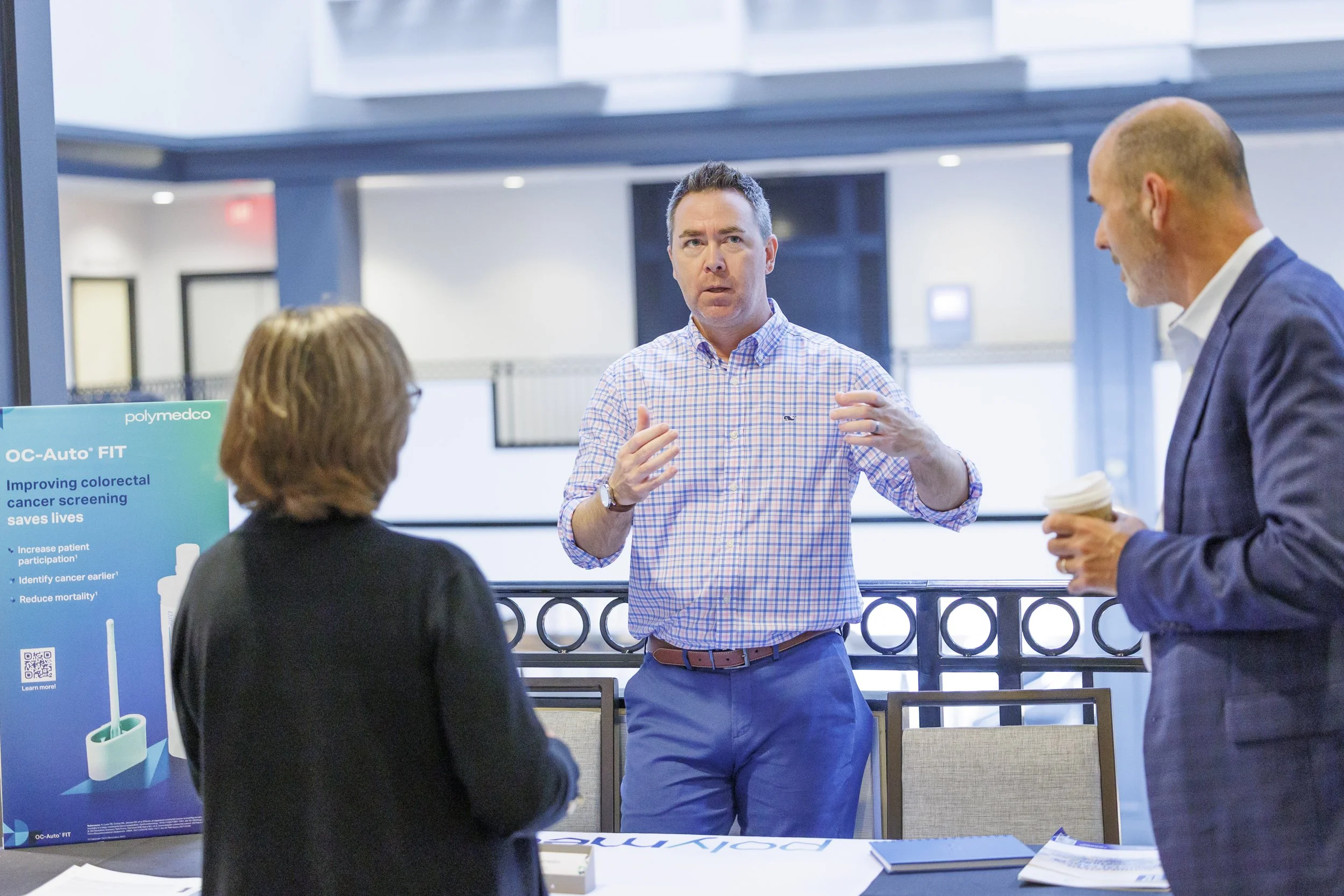 Three people are having a discussion in a conference or meeting room. One man is speaking, and two others are listening. There is a promotional stand about colon cancer screening on the left side.