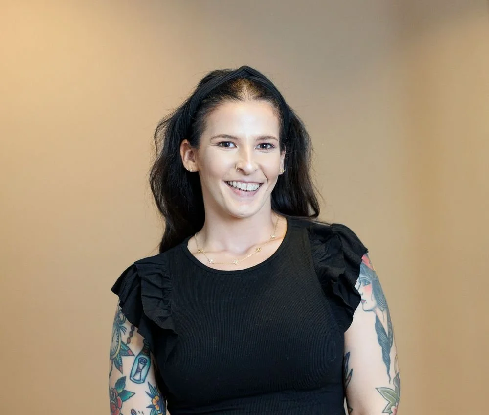 A woman with dark, wavy hair, wearing a black top with ruffled sleeves, smiling, with colorful tattoos on her arms, standing against a plain background.