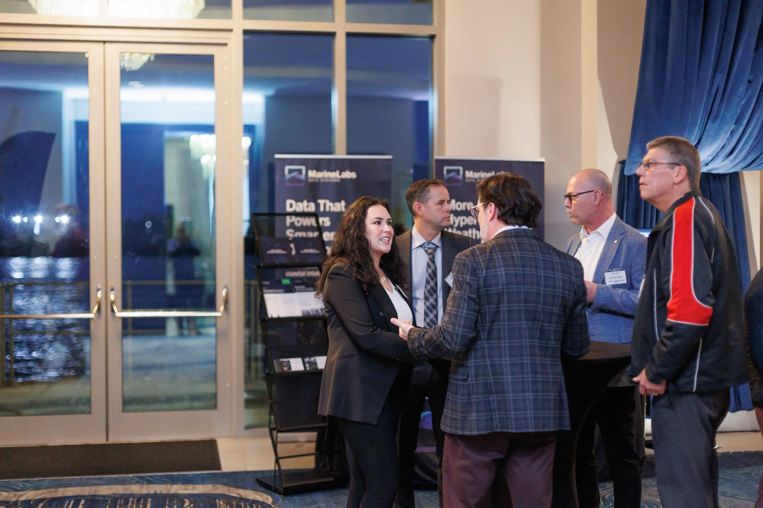 A group of five professionals engaged in conversation at a conference or networking event in an indoor venue with large windows and blue curtains, with conference banners in the background.