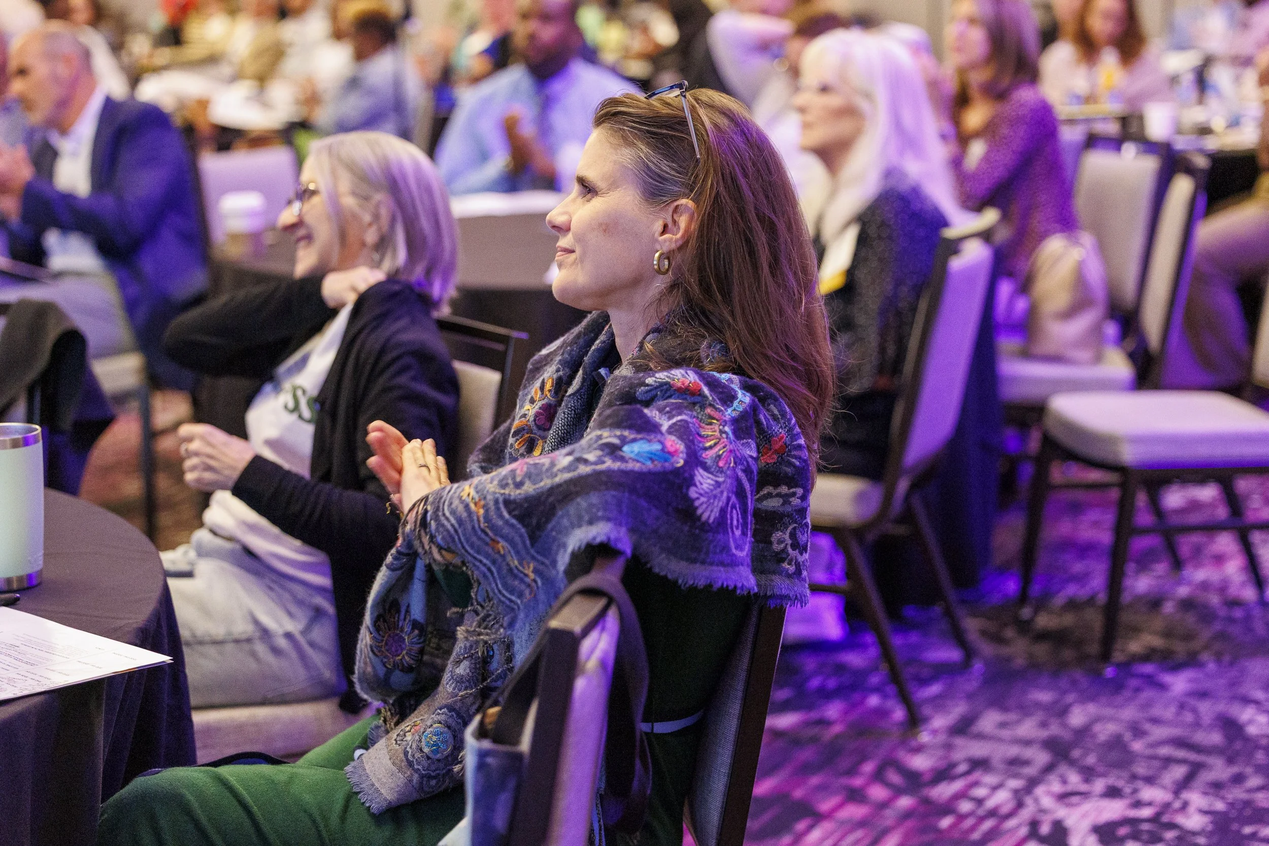 A woman listening attentively at a conference, seated at a table with other attendees in the background.