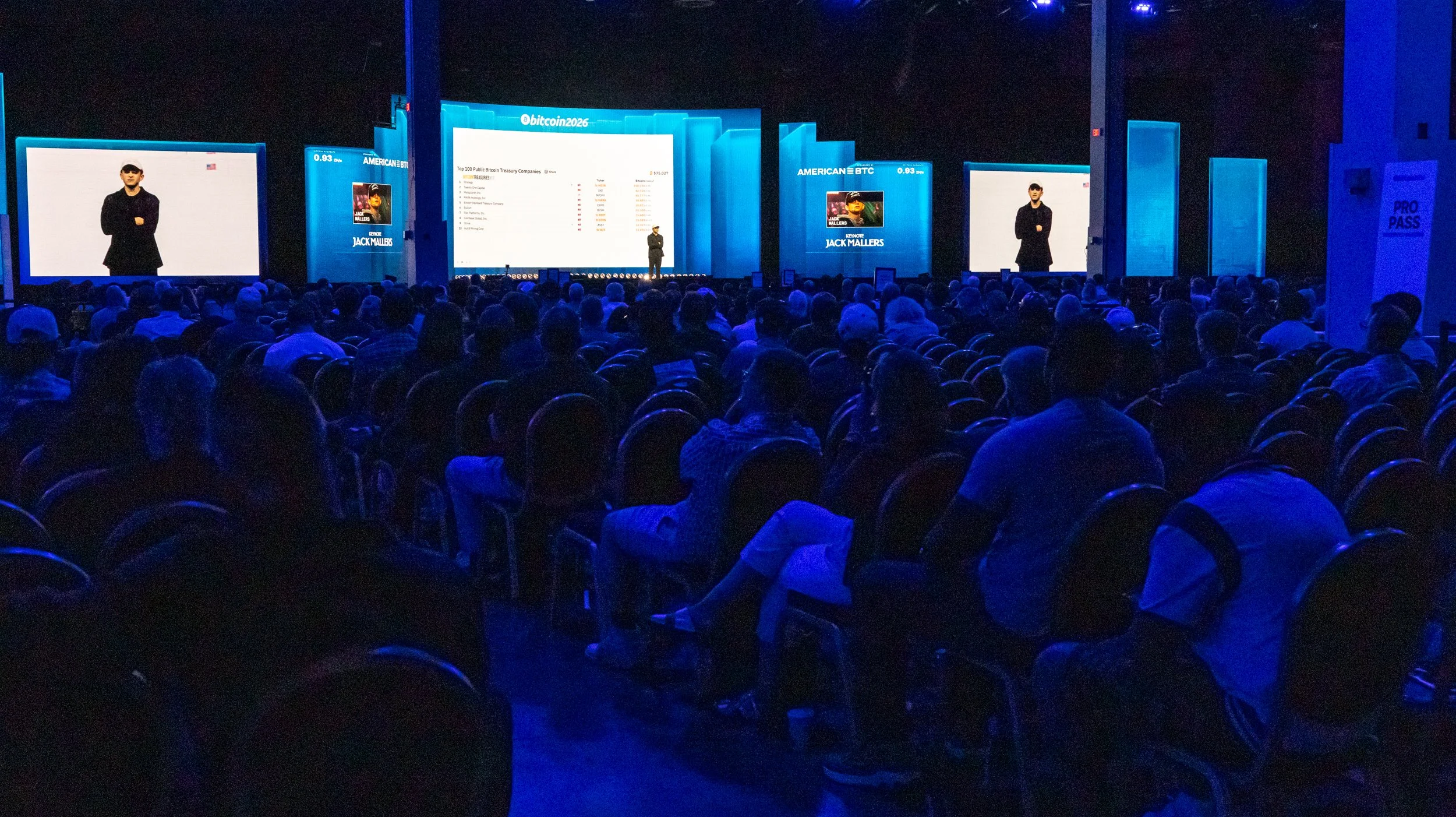 A large conference or presentation event with many attendees seated in rows, facing a stage with large screens displaying a speaker and presentation slides in a dark blue-lit room.