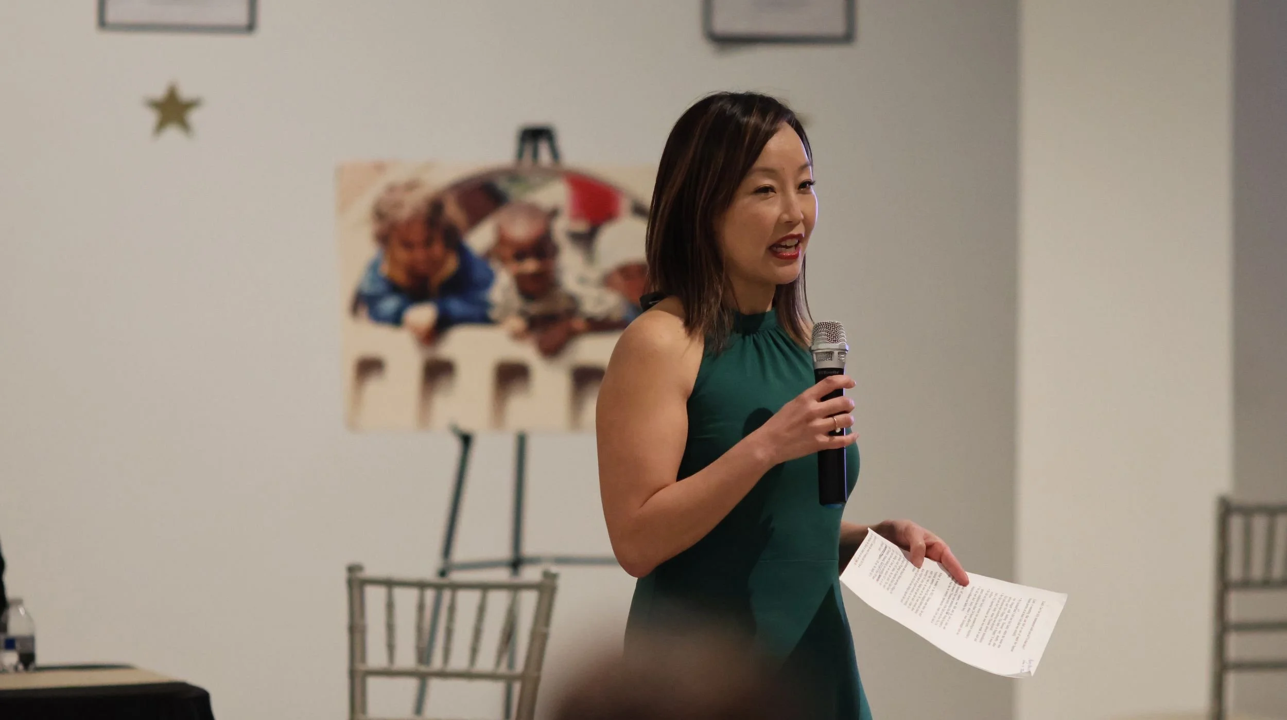 A woman in a green sleeveless dress speaks into a microphone while holding a paper, standing in front of a white wall with framed pictures and a blurred photograph of two children.