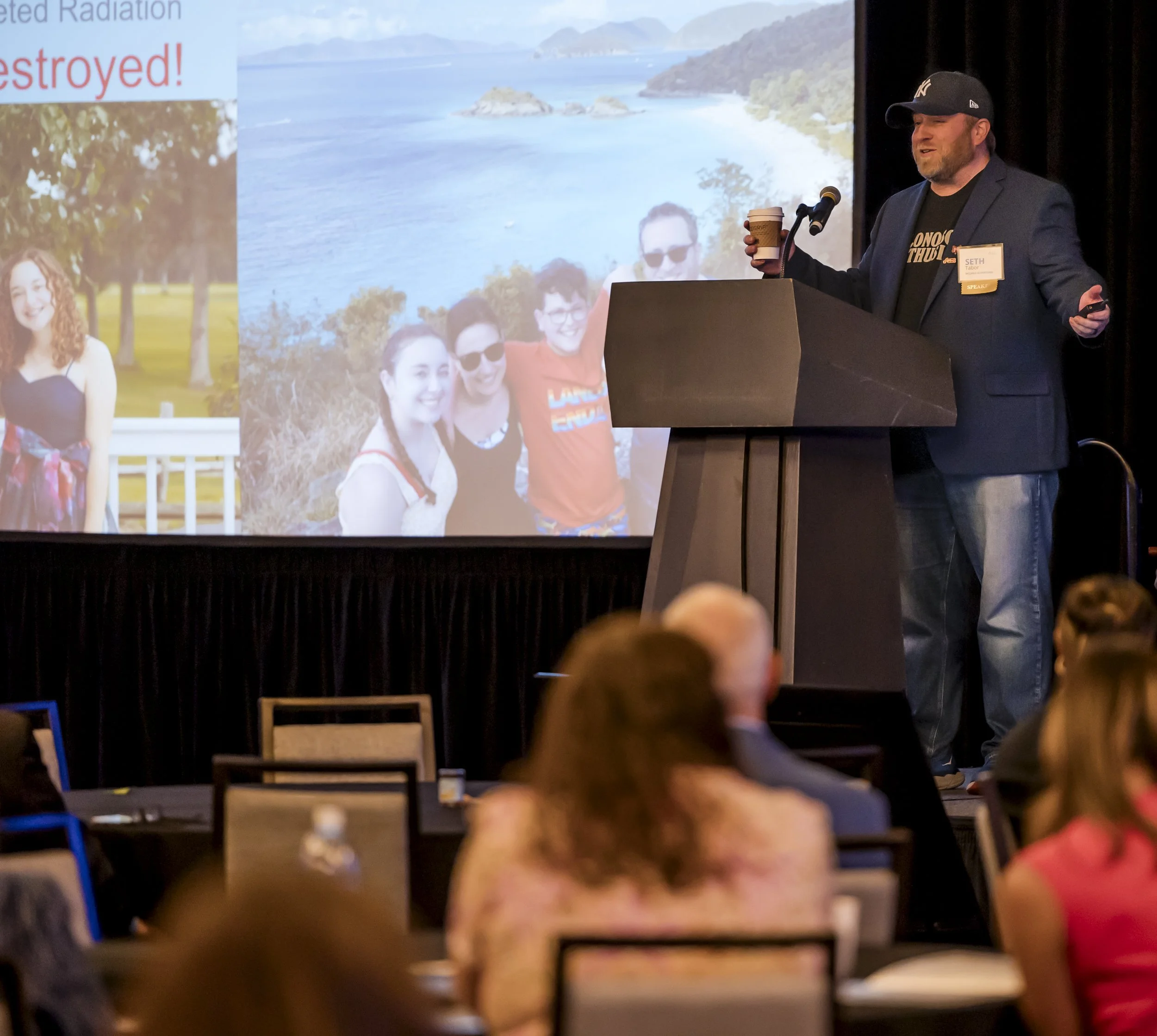 A man giving a presentation on a stage with a microphone and a takeaway coffee cup. The presentation slide behind him shows a photo of four people outdoors near a body of water, with a second photo of a woman in a park. Audience members are seated at