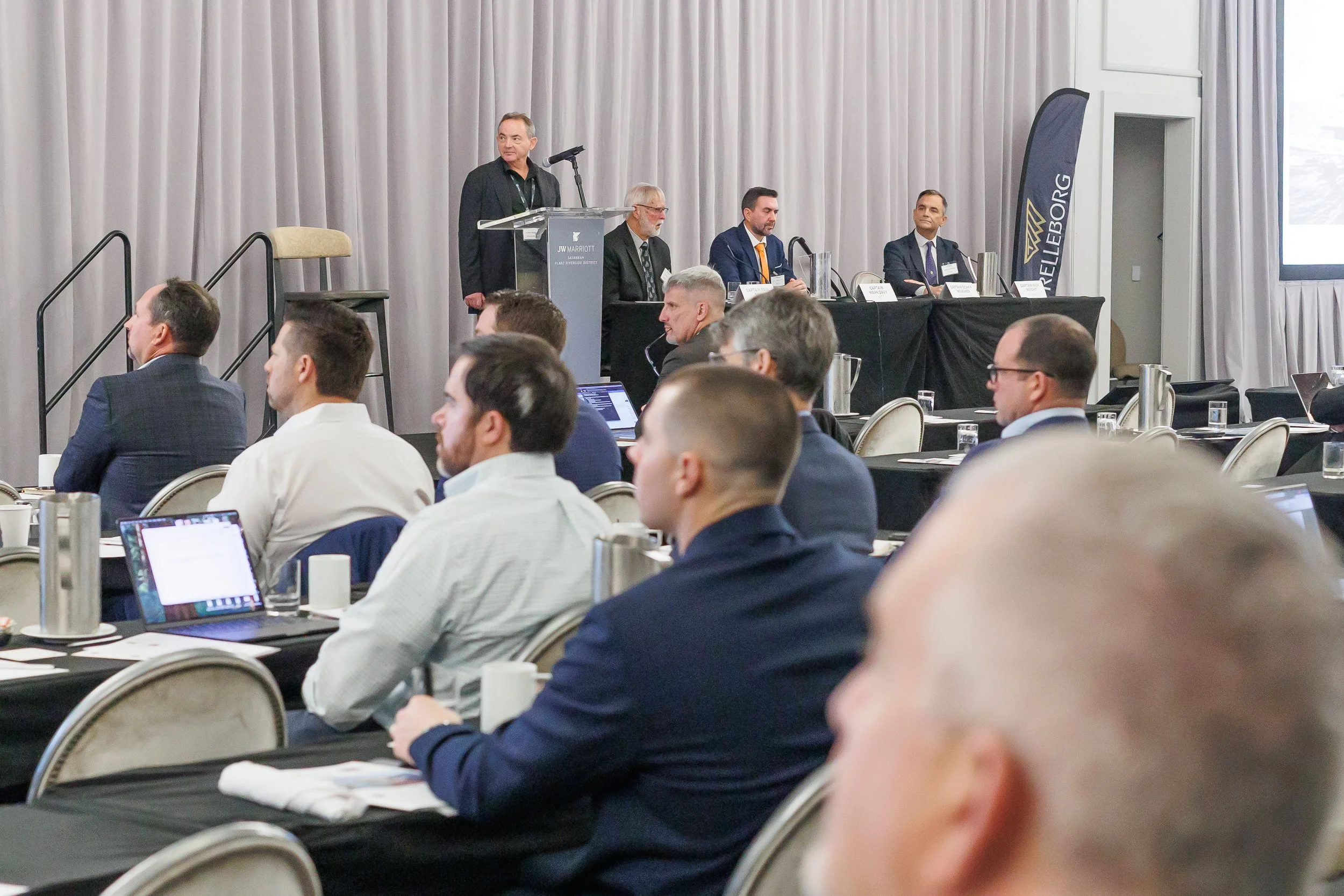 Business conference with panel of speakers at podium, audience taking notes and listening in conference room.
