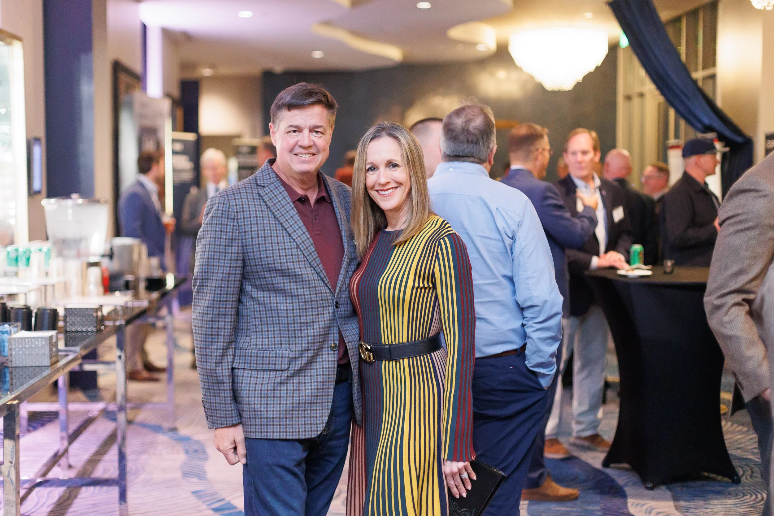 A man and woman standing together at a professional social event, smiling at the camera, with other attendees and a bar in the background.
