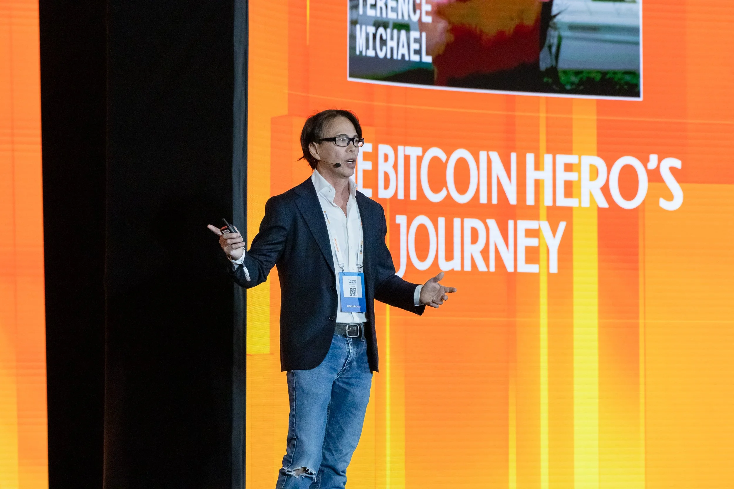 A man with glasses and a microphone headset giving a presentation on stage. Behind him is a large orange and red screen with the text 'CONFERENCE MICHAEL' and 'CRYPTOCURRENCY'S JOURNEY'.