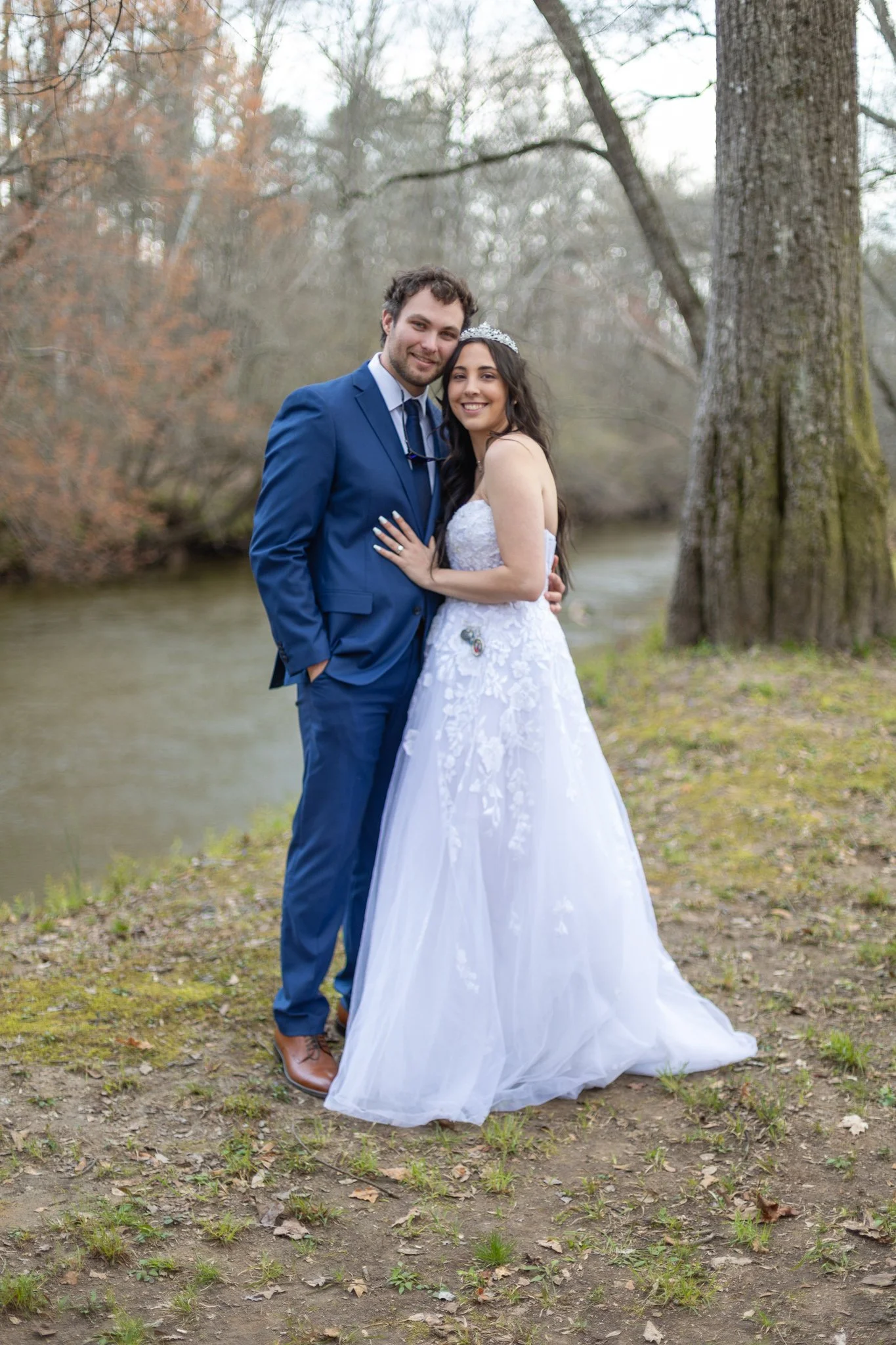 A newlywed couple with the groom in a blue suit and the bride in a white wedding dress standing outdoors near a river, embracing each other and smiling, with trees in the background.