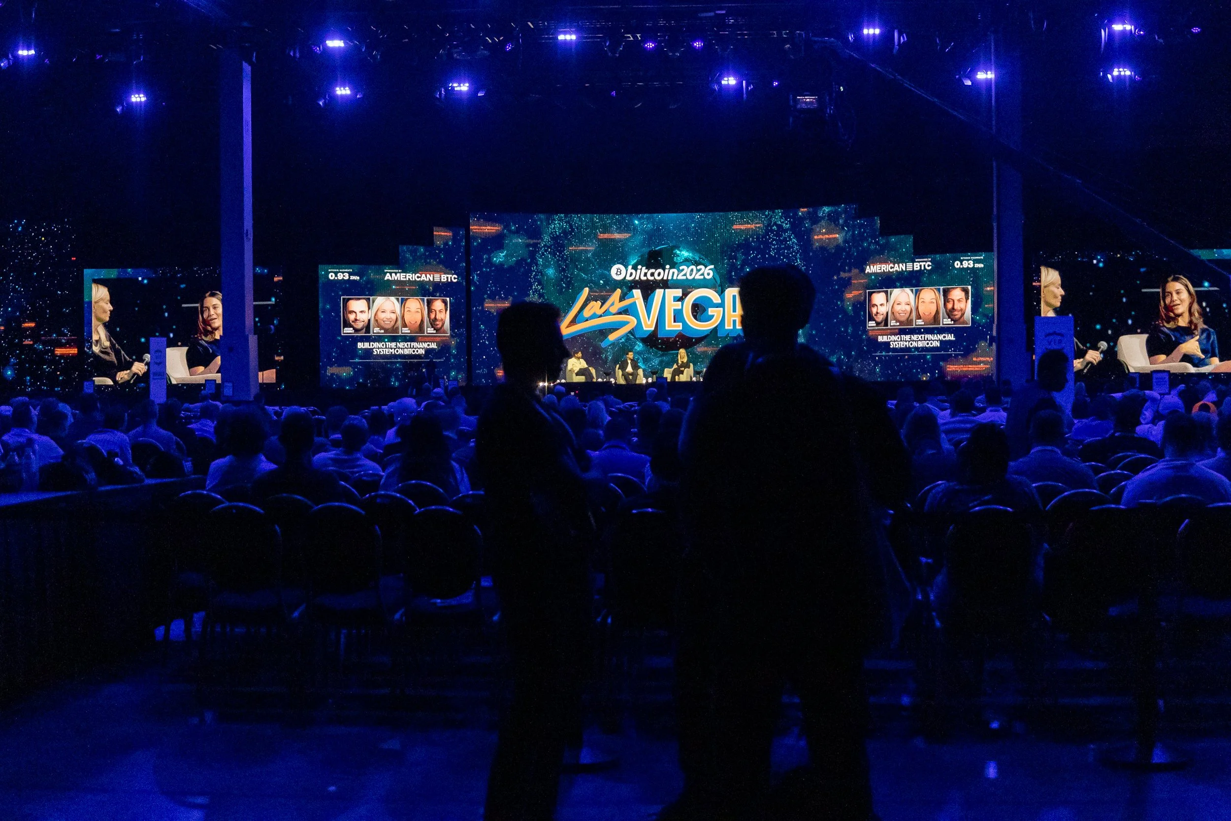 Dark indoor stage with large screen displaying 'Bitcoin 2026 Las Vegas' and lineups of speakers, with people seated in front and some standing, illuminated by blue and purple lights.