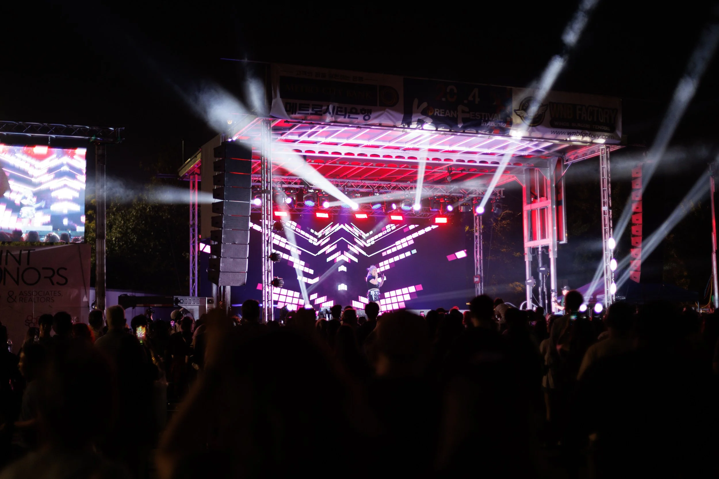 Nighttime outdoor concert stage with vibrant purple and pink lighting, large LED screens, and a crowd of people watching.