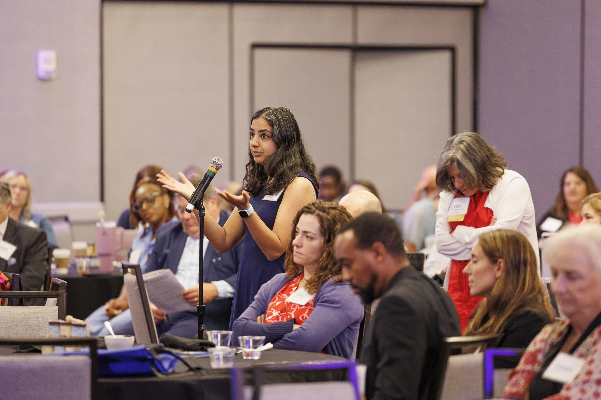 A woman standing at a microphone asking a question during a conference or meeting, with audience members seated and listening.