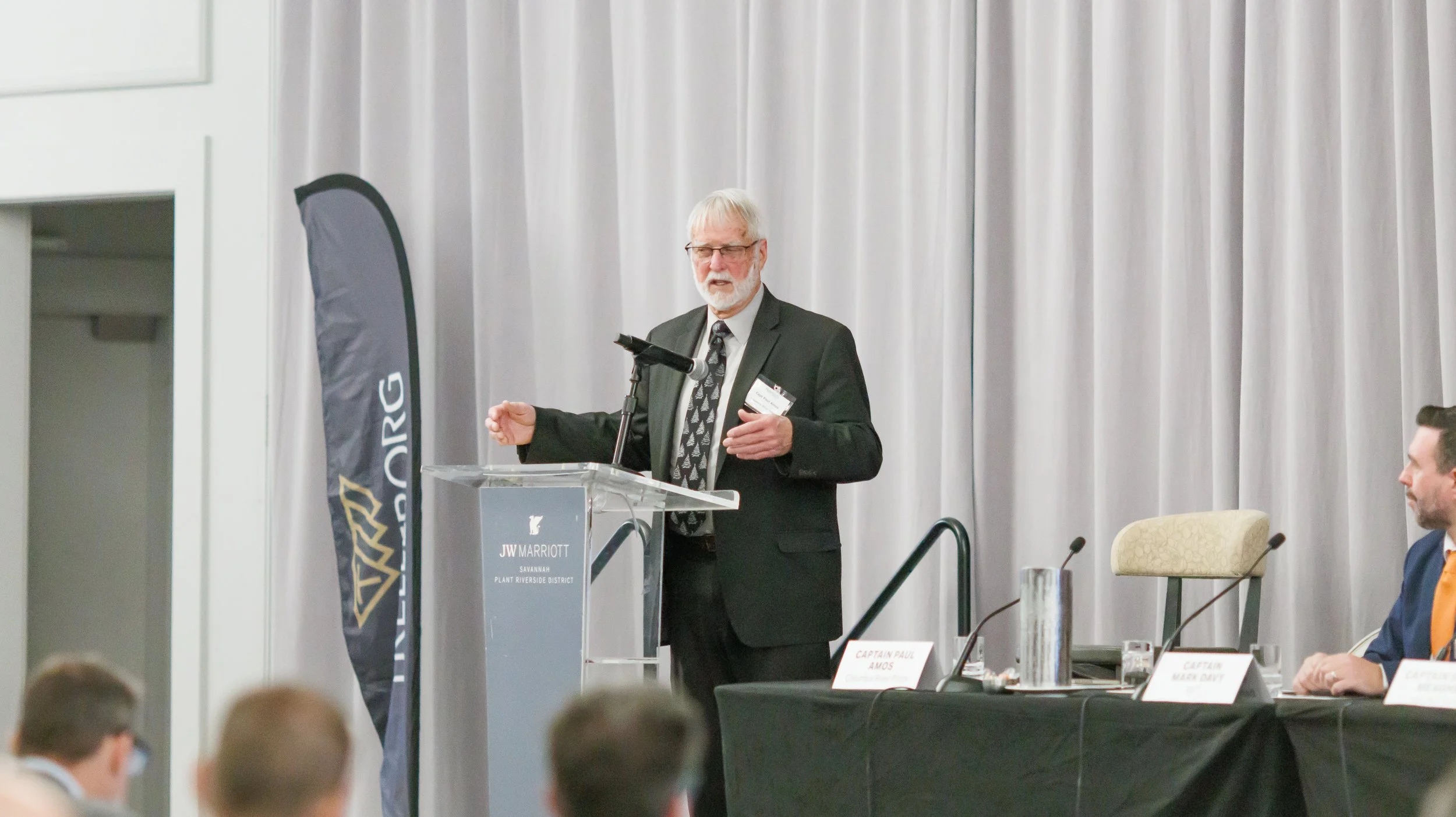An elderly man with white hair and beard, wearing glasses and a black suit, speaks at a podium during a conference held at JW Marriott in Savannah, Plant Riverside District. He is gesturing with his right hand, and a microphone is positioned in front