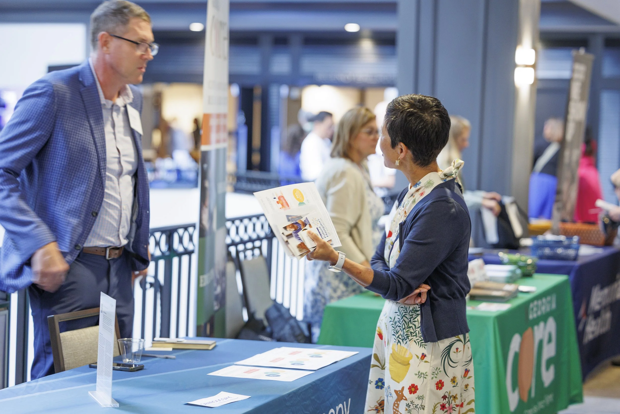 A woman in a dark jacket and floral dress is speaking with a man in a blue checkered suit at an indoor event. The woman is holding brochures and gesturing as she talks. Several tables with informational materials and banners are visible in the backgr