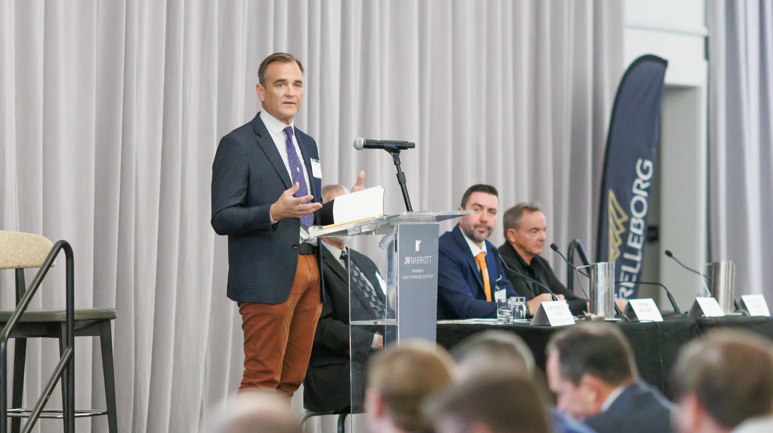 Man in a suit speaking at a podium during a conference or panel discussion, with three other men sitting at a table behind him and a black banner with blue and white text on the right side of the image.