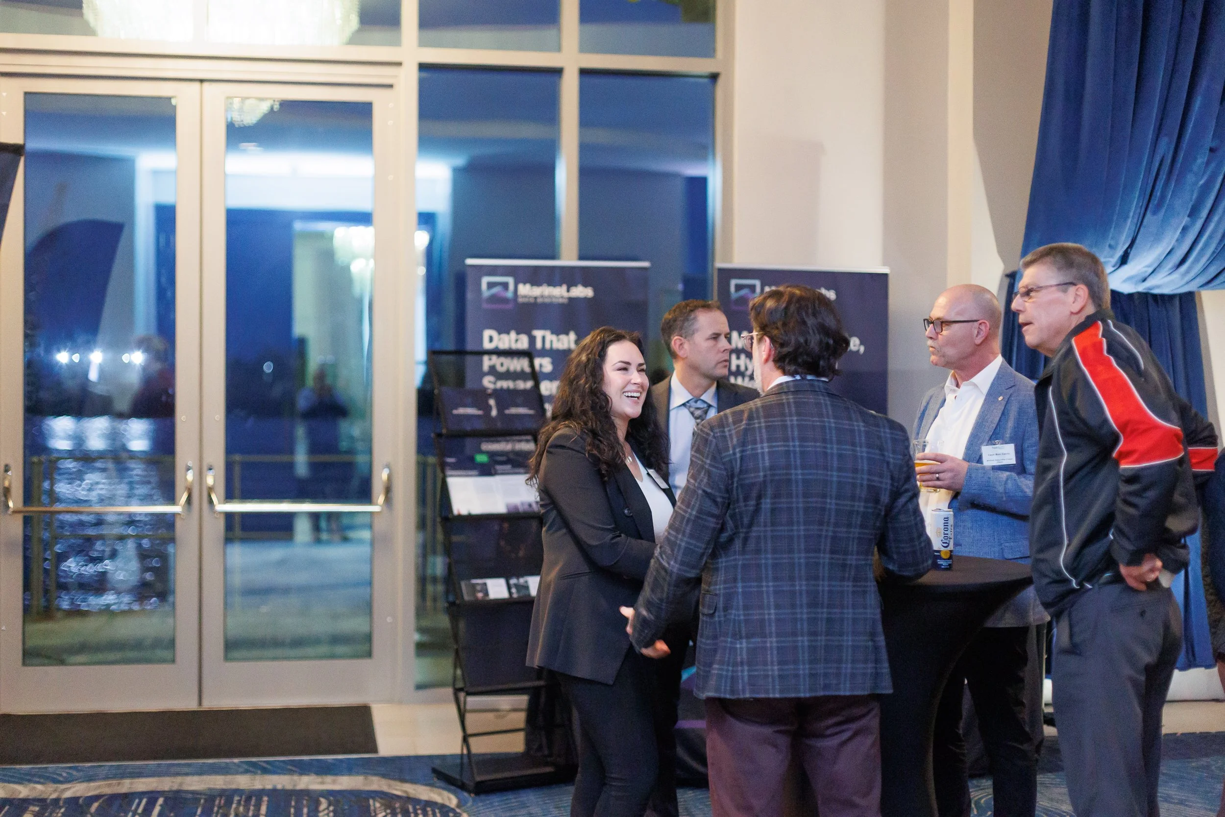 Group of five professionals engaging in conversation at a networking event, with promotional banners in the background, indoors at night.