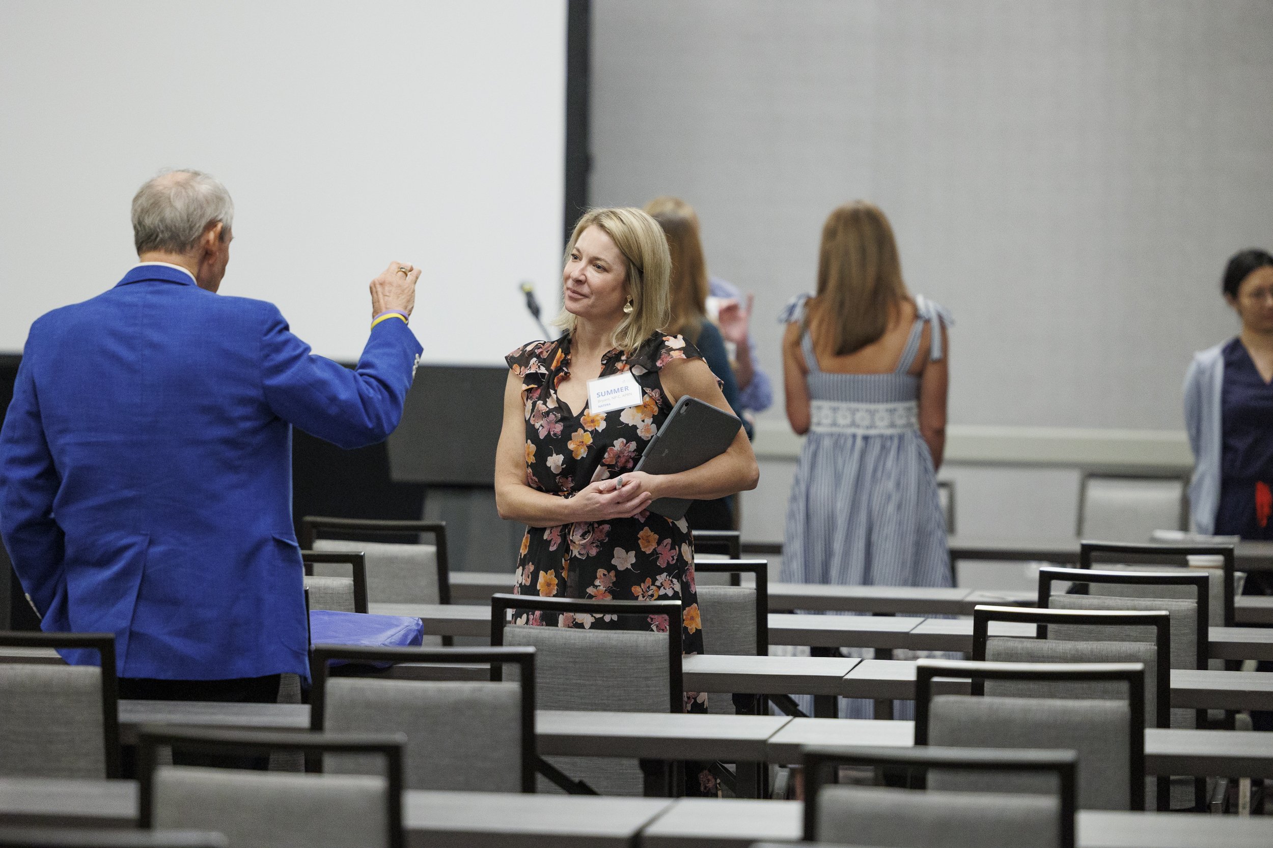 A woman in a floral dress holding a folder talking to an older man in a blue suit in a conference room