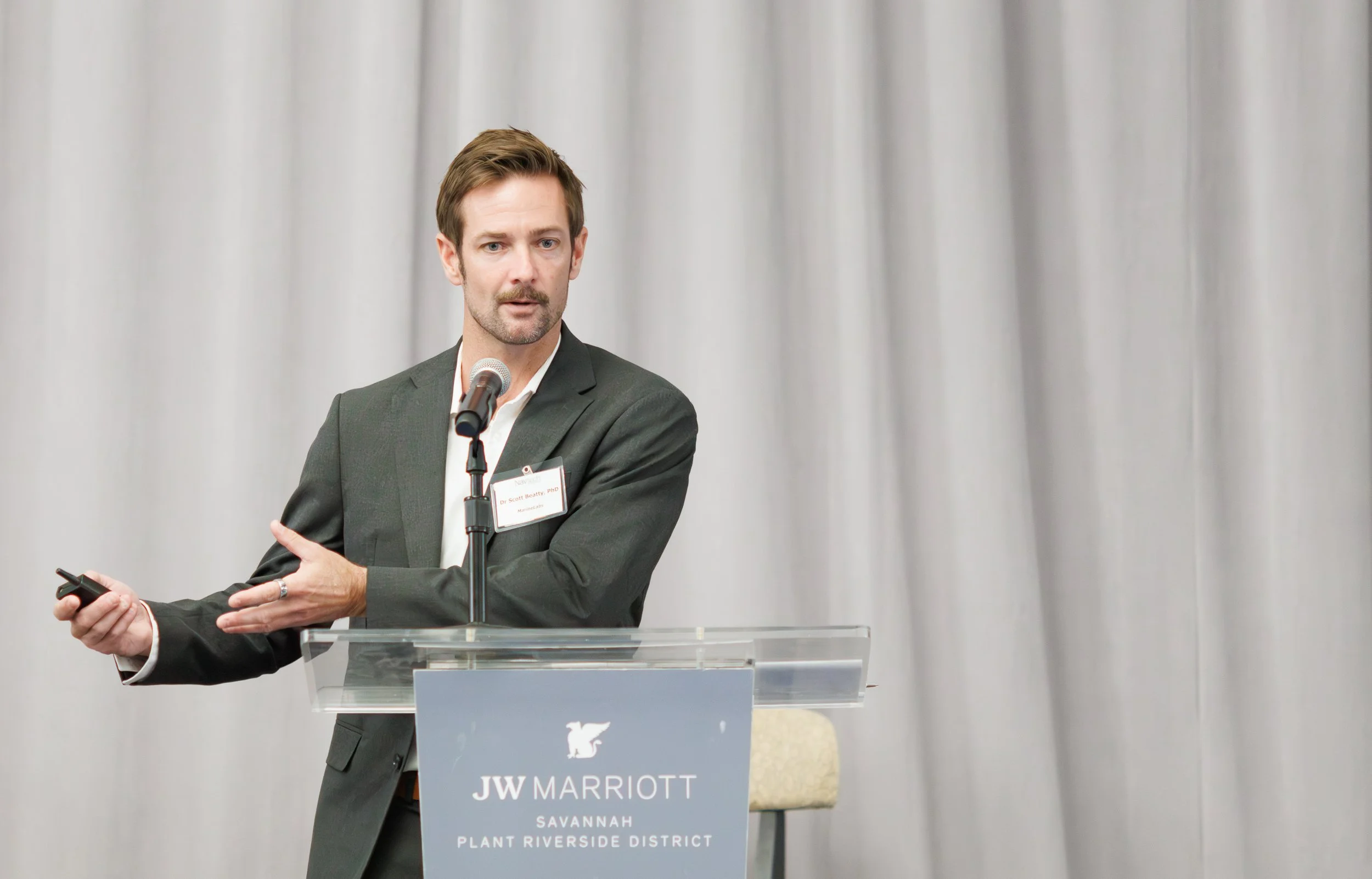 A man giving a presentation at a JW Marriott event, standing behind a podium with a microphone, gesturing with his right hand, wearing a dark suit and holding a clicker in his left hand, with a gray curtain background.