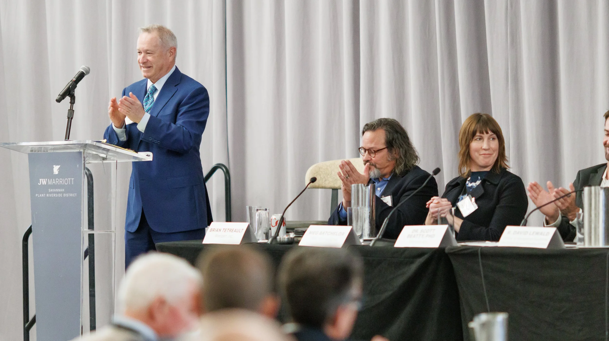 A man in a blue suit standing at a podium with a microphone, smiling and clapping. Four other individuals are seated at a table behind him, clapping and smiling, with name placards in front of them. The setting appears to be a conference or panel dis