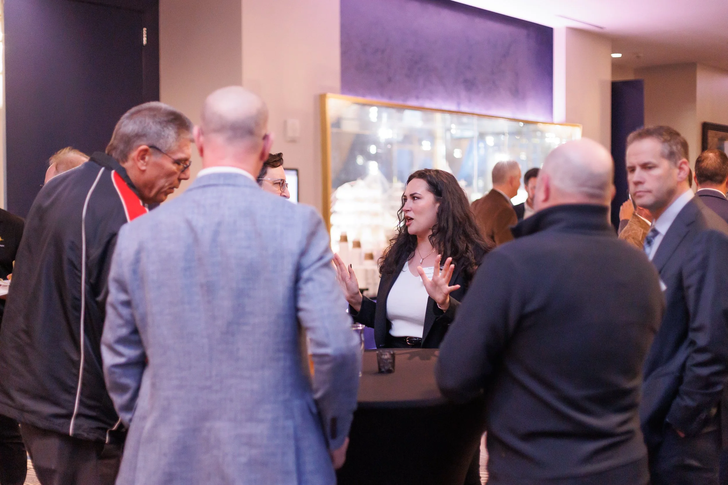 A woman in a black blazer and white top speaking to a group of men at a networking event or conference in an indoor venue.