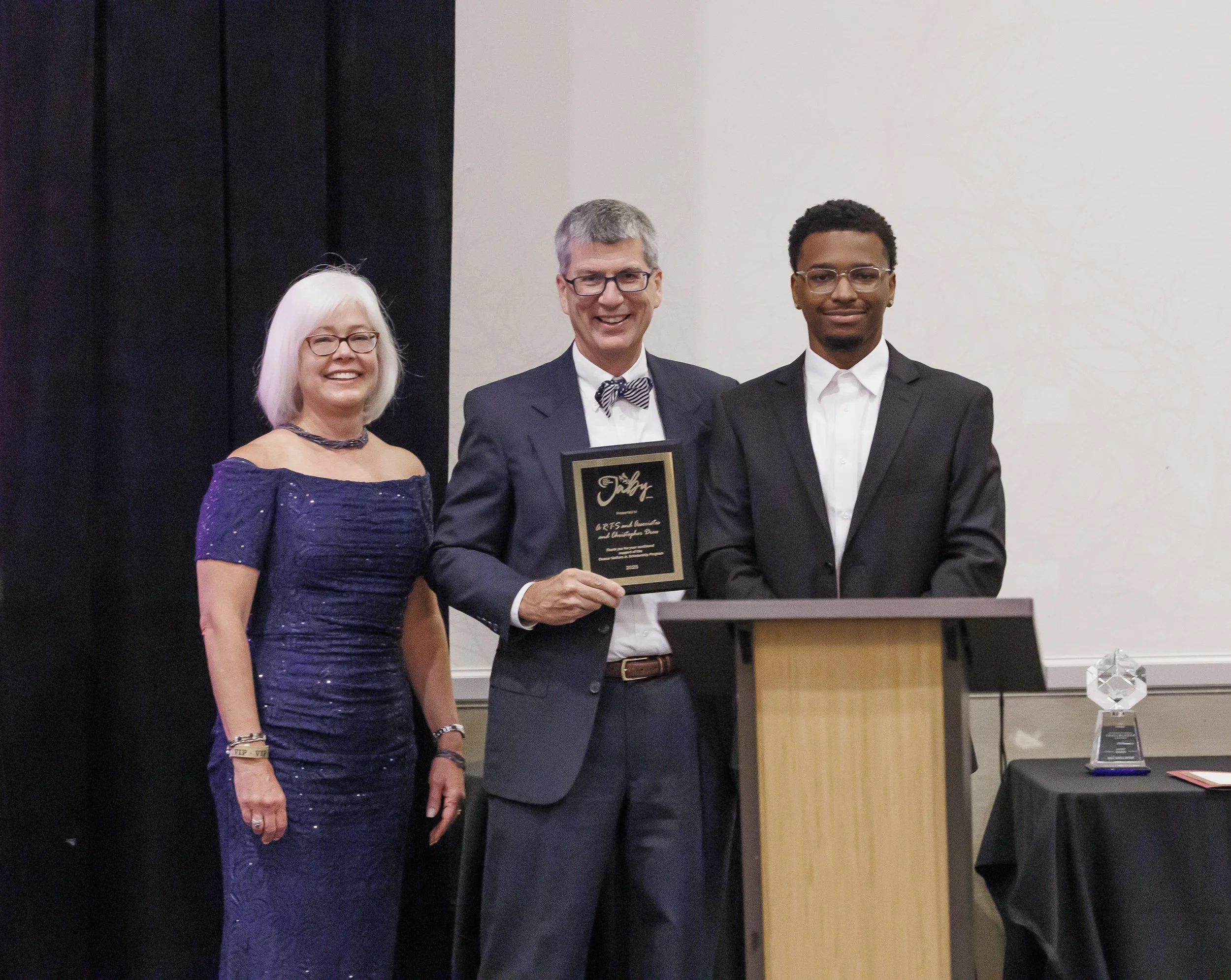 Three people dressed in formal attire standing together at an awards ceremony, with a man holding a plaque.
