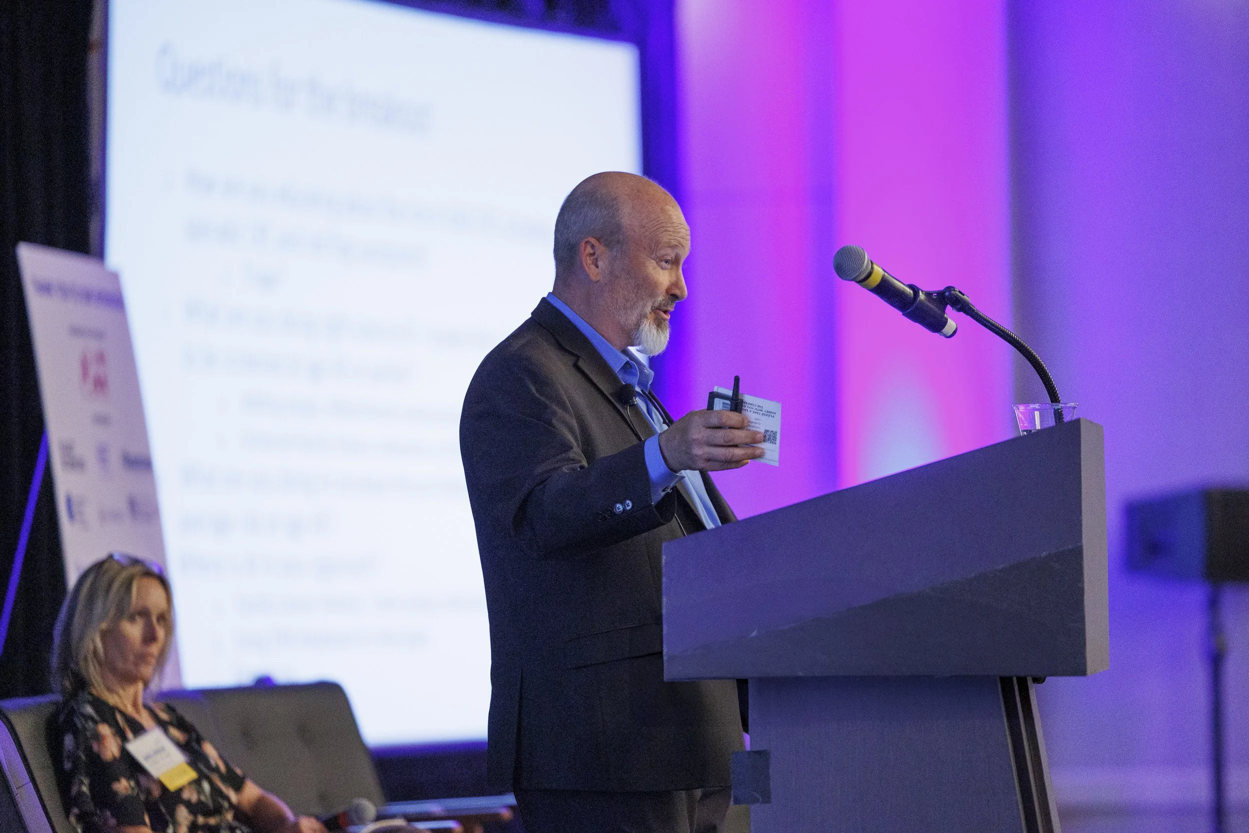 A man in a suit giving a presentation at a conference, holding a microphone and a badge, with a large screen and a woman sitting in the background.