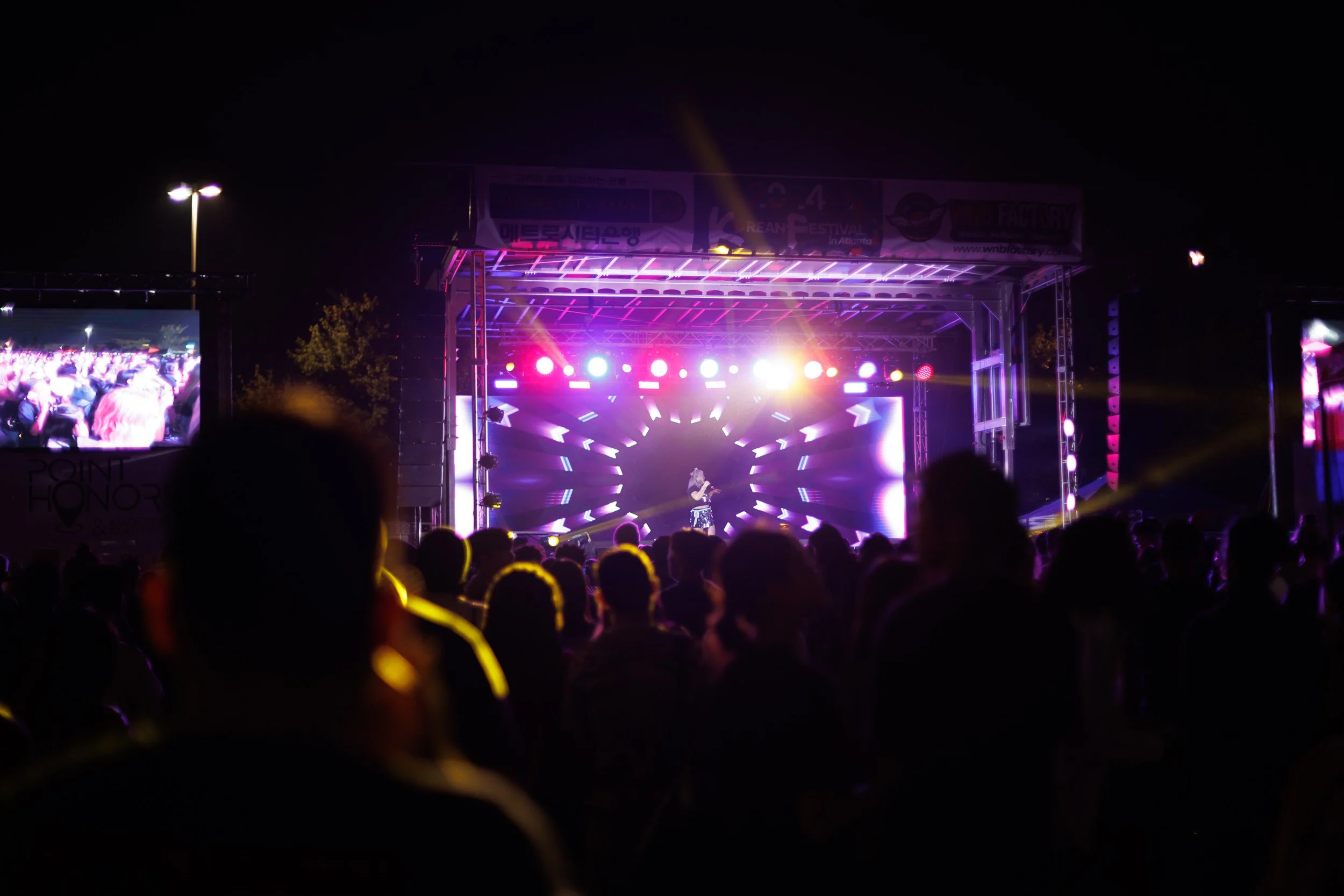 Nighttime outdoor concert stage illuminated with colorful lights, surrounded by an audience.