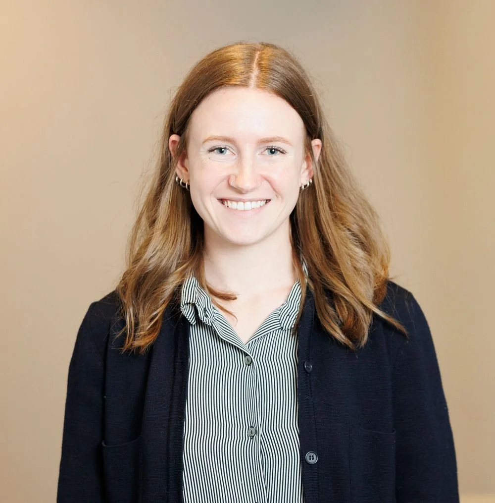 A young woman with shoulder-length red hair, wearing a black cardigan over a collared striped shirt, smiling at the camera against a beige background.